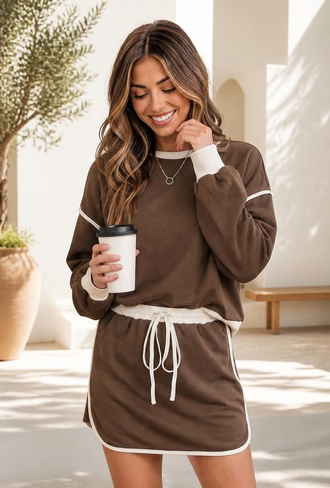 A woman wearing a mocha brown sweatshirt and mini skort set with cream contrast trim, holding a coffee cup.