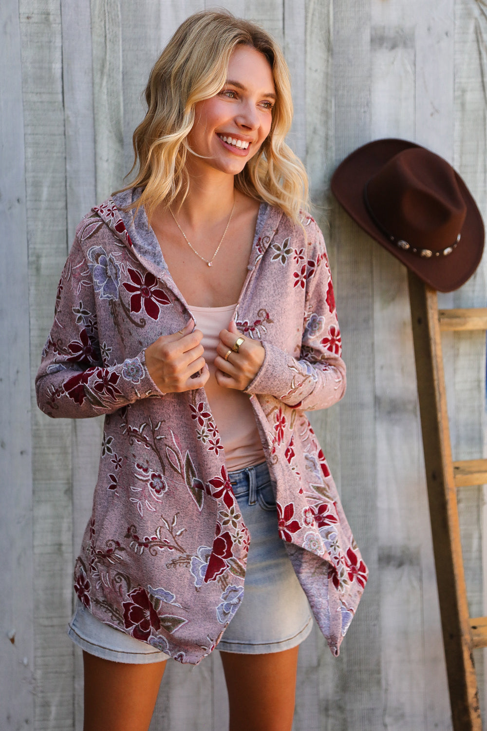 Woman wearing a floral cardigan in front of a wooden wall with a hat and ladder.