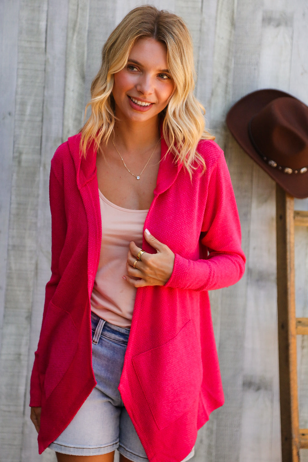 Woman wearing a bright pink cardigan over a light pink top and denim shorts, standing against a wooden wall.