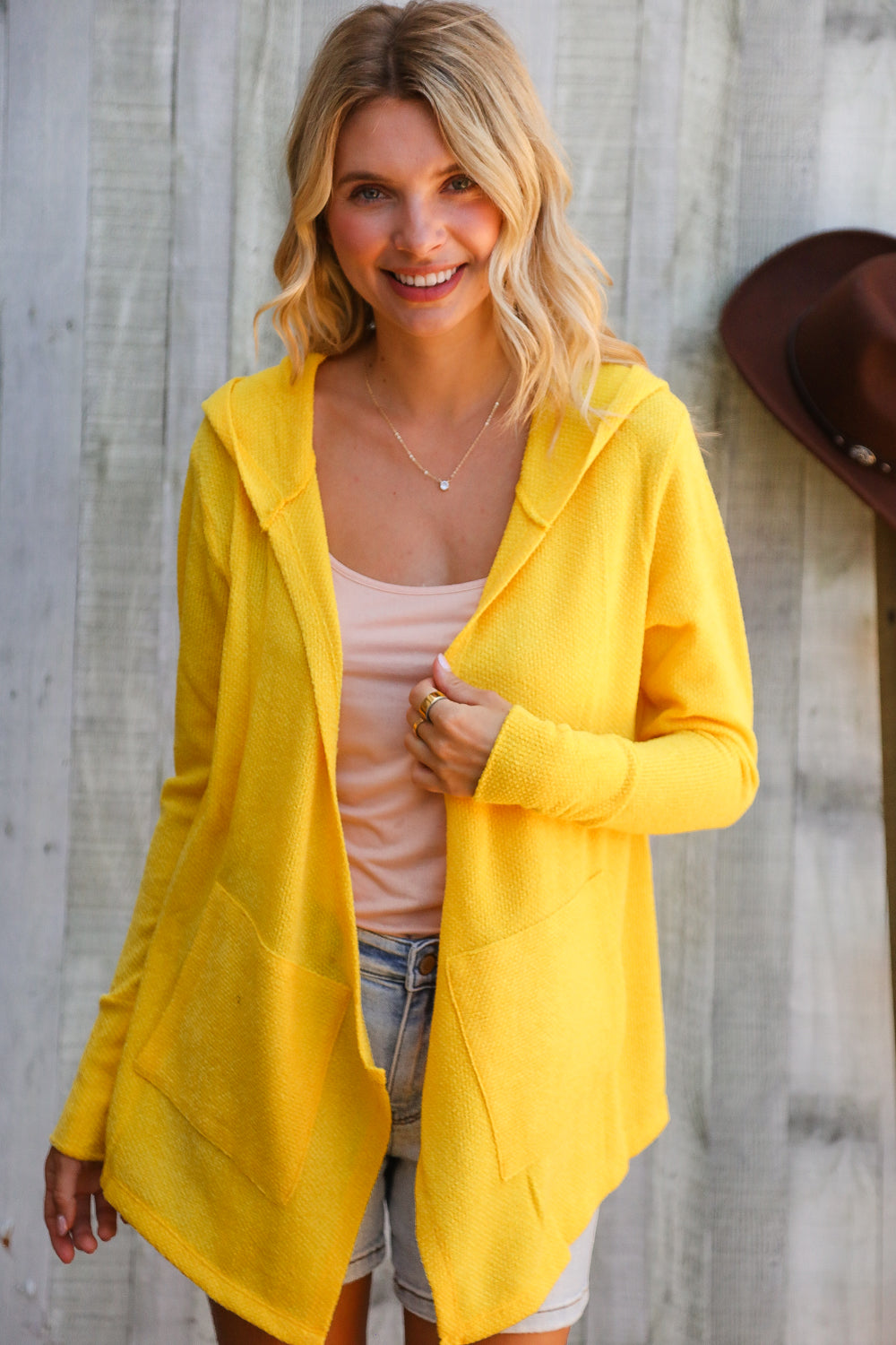 Woman wearing a yellow cardigan over a pink top and denim shorts, standing against a wooden wall.