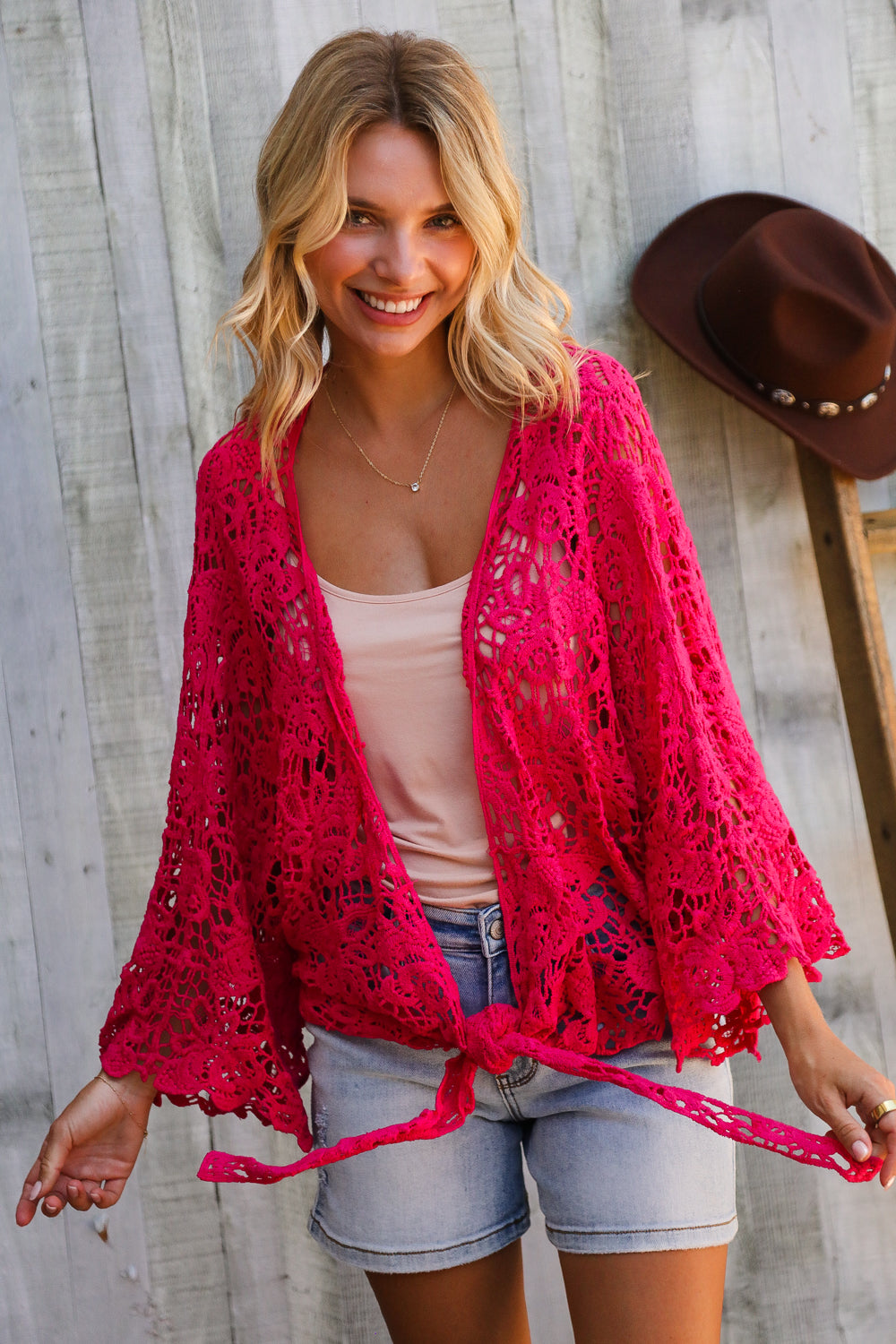 Woman wearing a bright pink crochet cardigan over a white top and denim shorts, standing against a wooden wall with a cowboy hat.