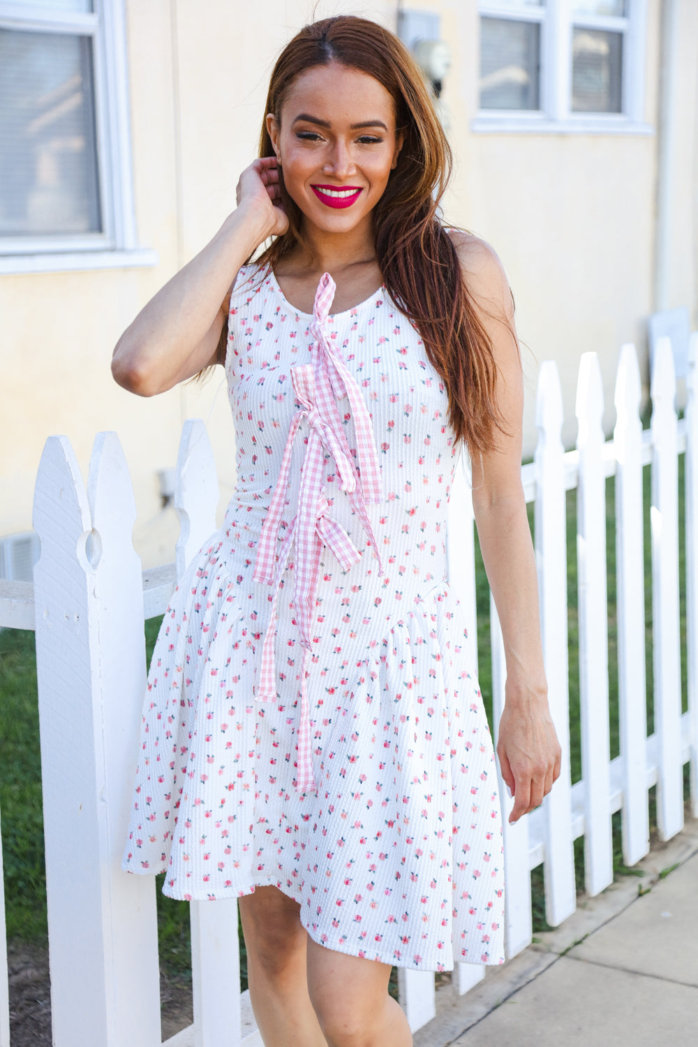Woman wearing a white floral dress with a pink ribbon in front of a white picket fence.