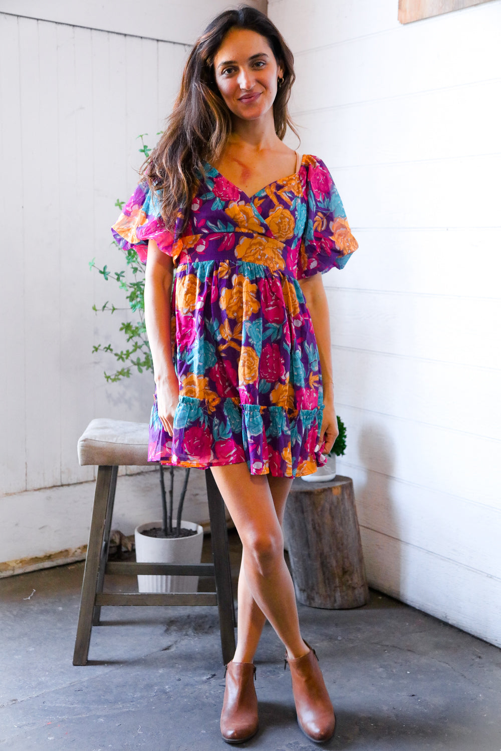 Woman wearing a colorful floral dress standing in a room with a white wall and wooden stool.