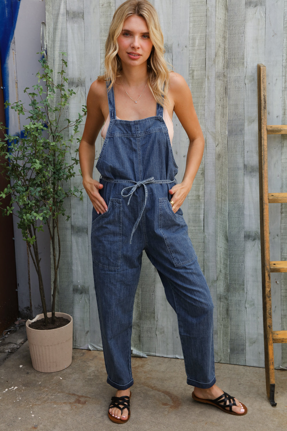 Woman wearing a denim jumpsuit standing against a wooden wall.