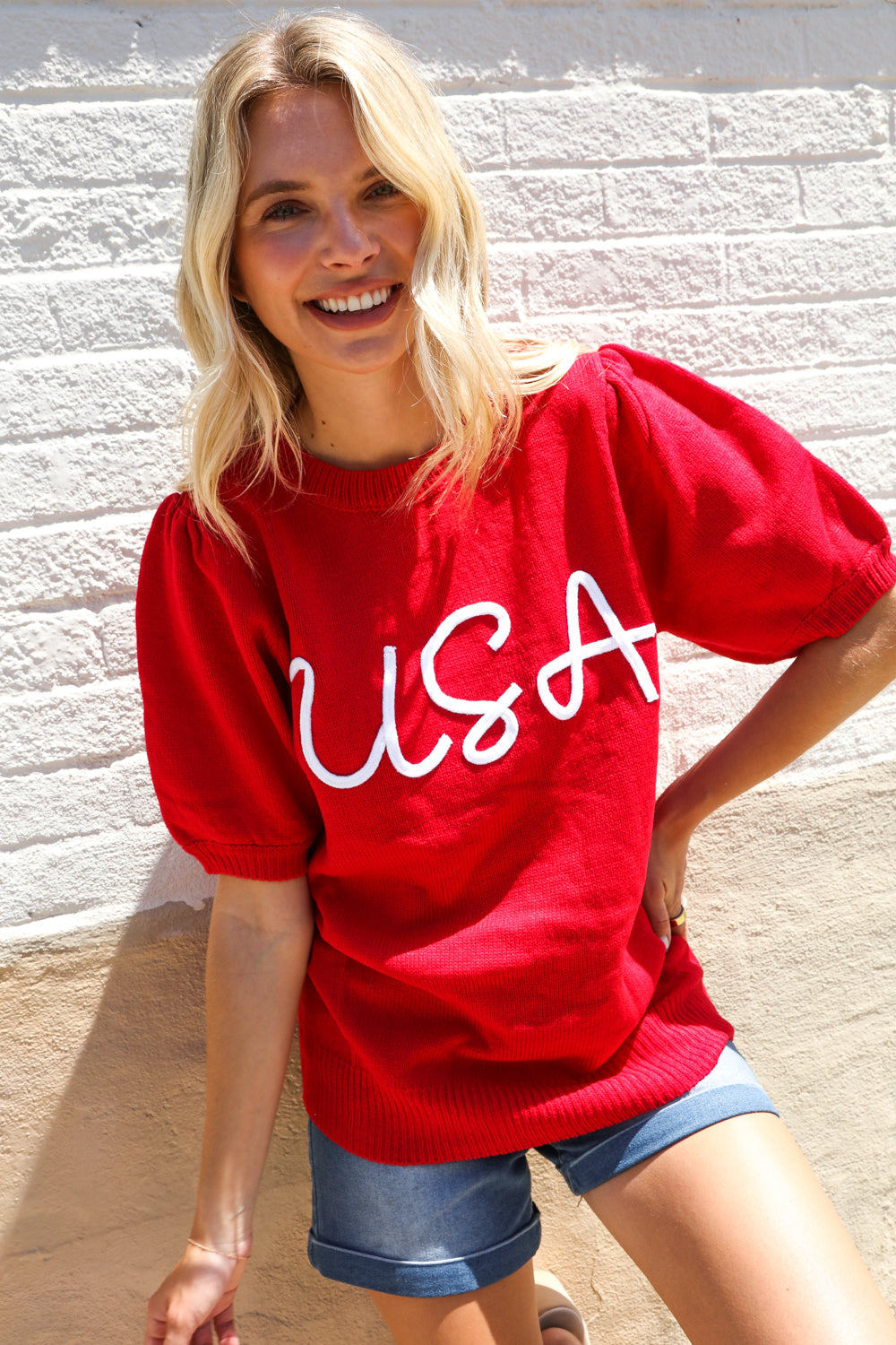 Woman wearing a red 'USA' t-shirt against a light stone wall.