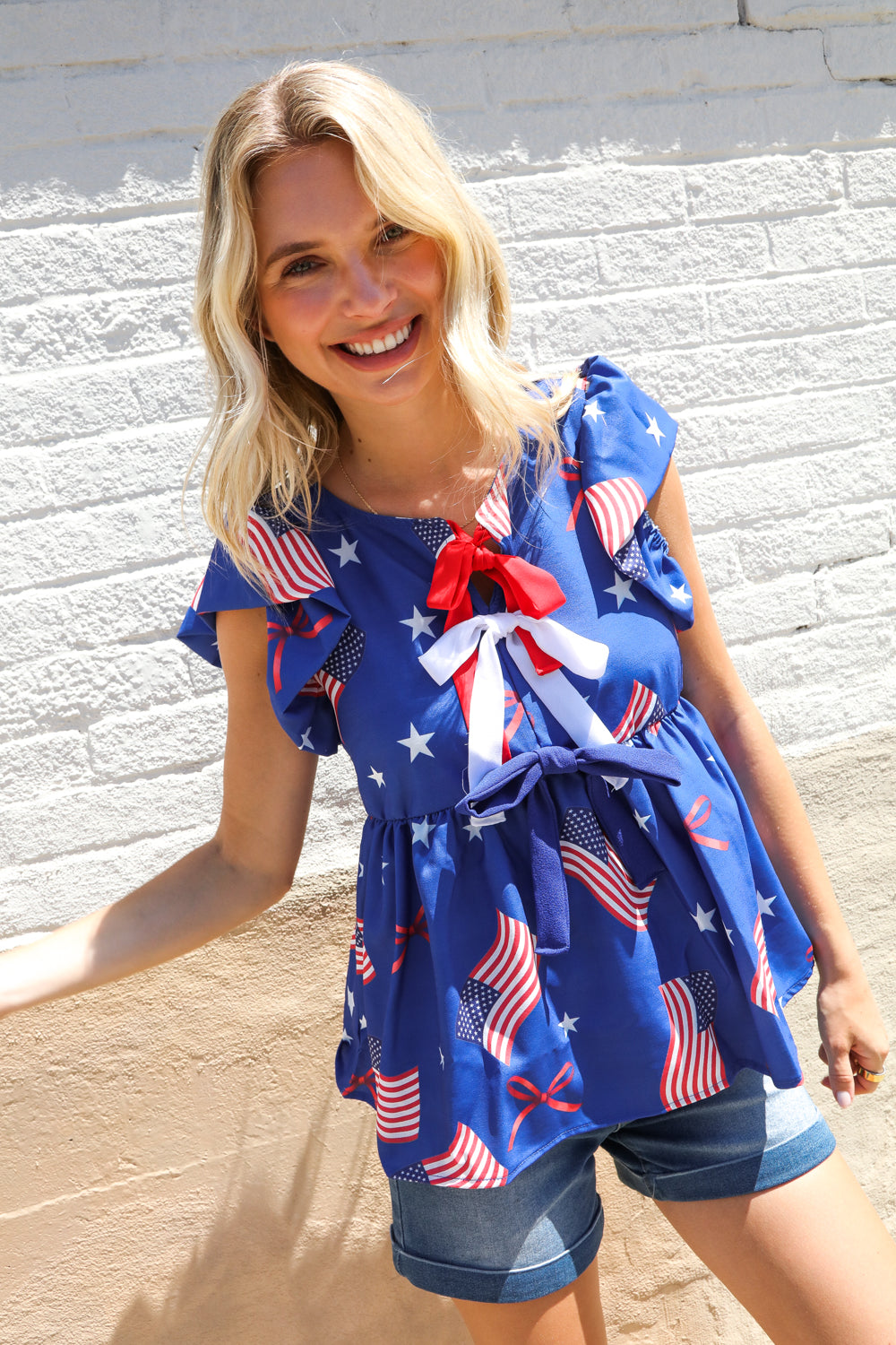 Woman wearing American Flag Blue & Red Stars & Bow Print Woven shirt Top against a light stone wall.