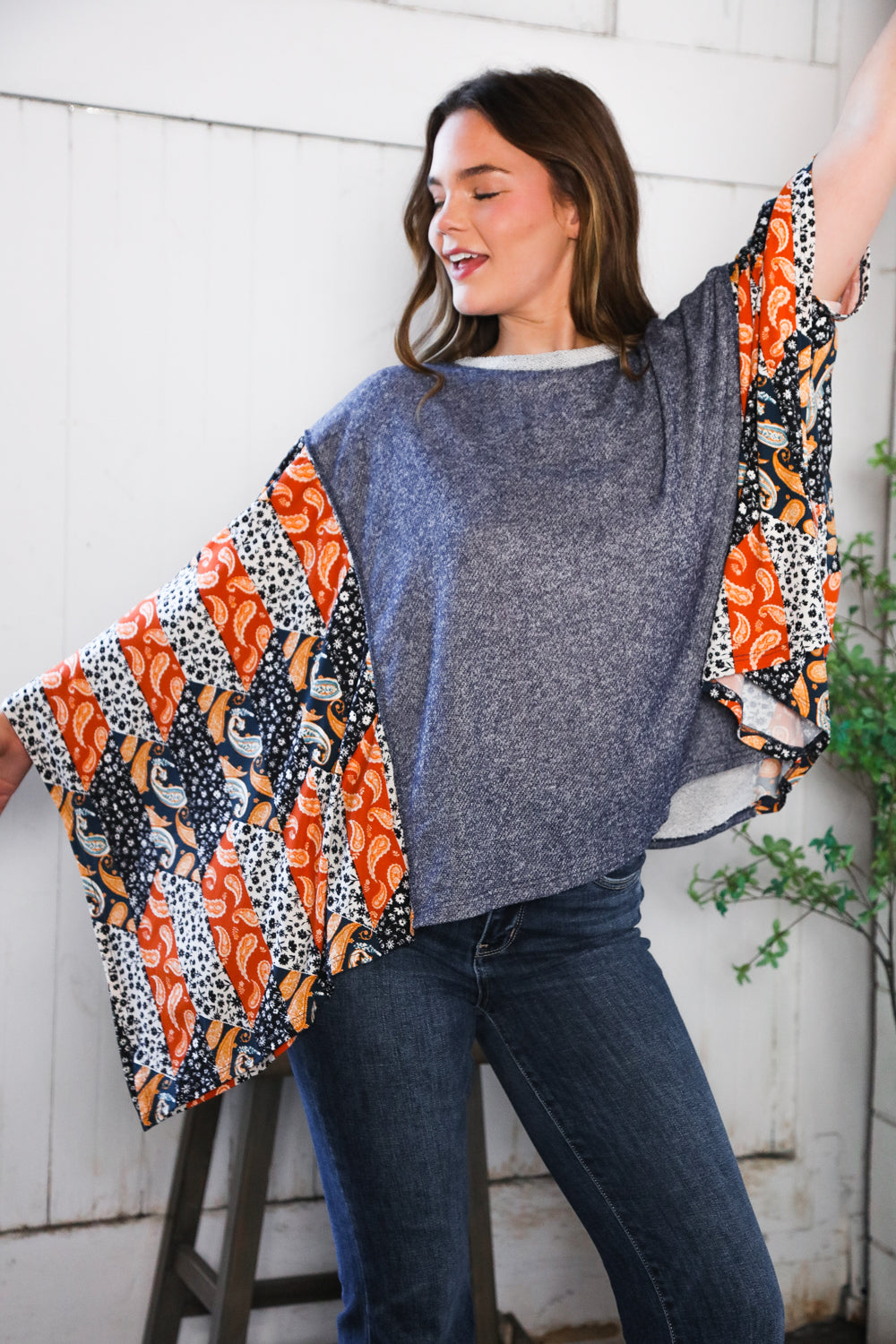 Woman wearing a gray French Terry Poncho Top with colorful sleeves against a white brick wall.