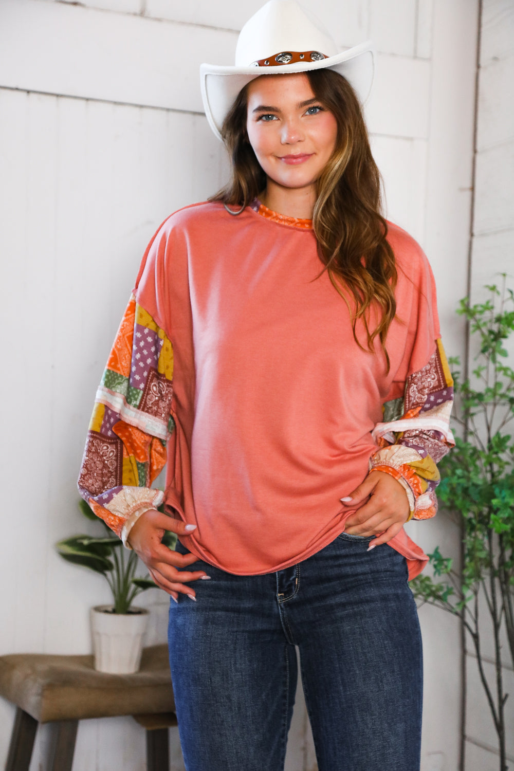 Woman wearing a colorful long-sleeve top and jeans with a white cowboy hat indoors.