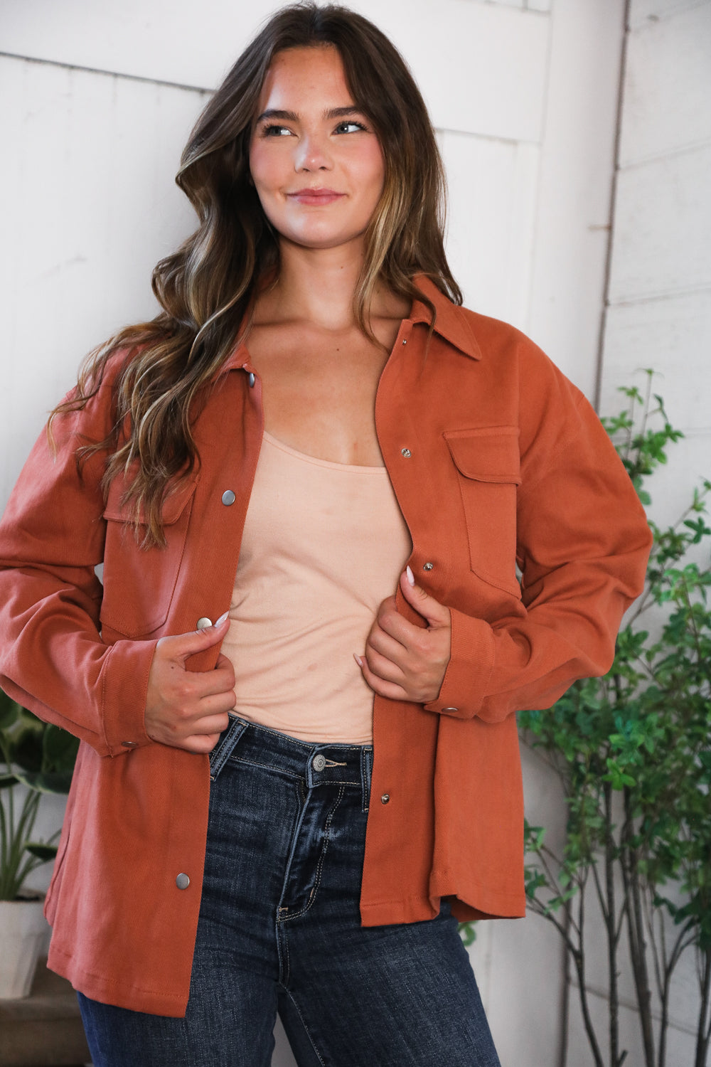 Woman wearing a Burnt Orange jacket over a beige top and blue jeans, standing against a white wall with plants.