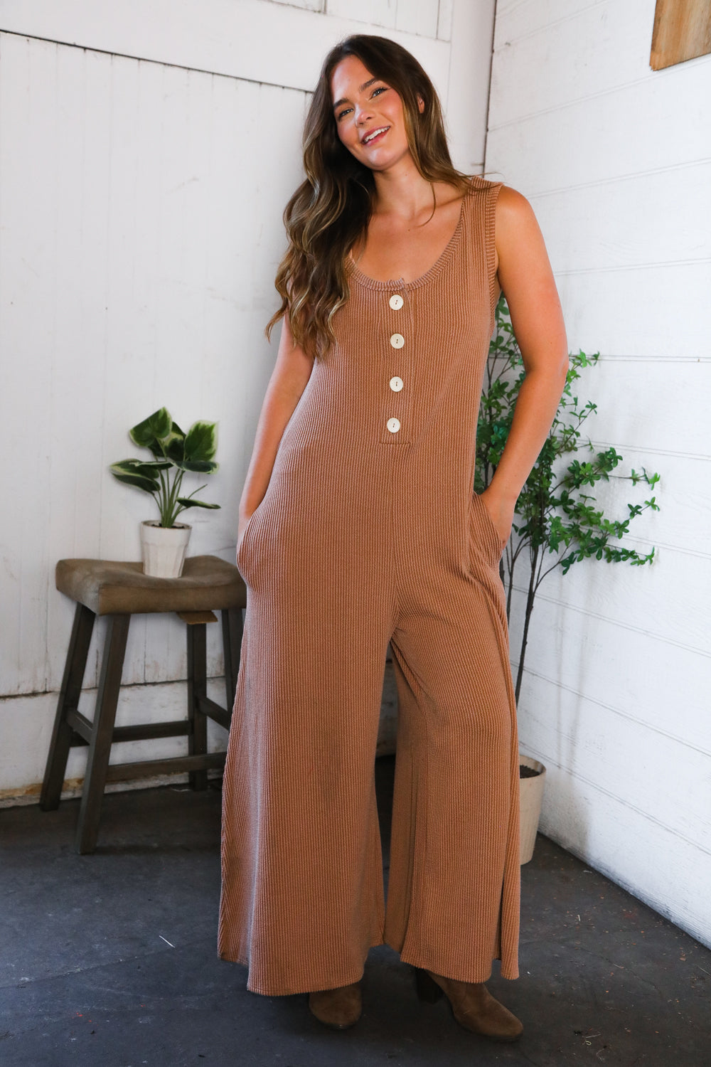 Woman wearing a brown jumpsuit standing in a room with white walls and plants.