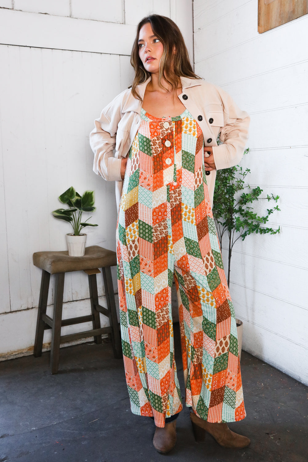 Woman wearing a colorful patchwork jumpsuit standing indoors with plants and a stool in the background.