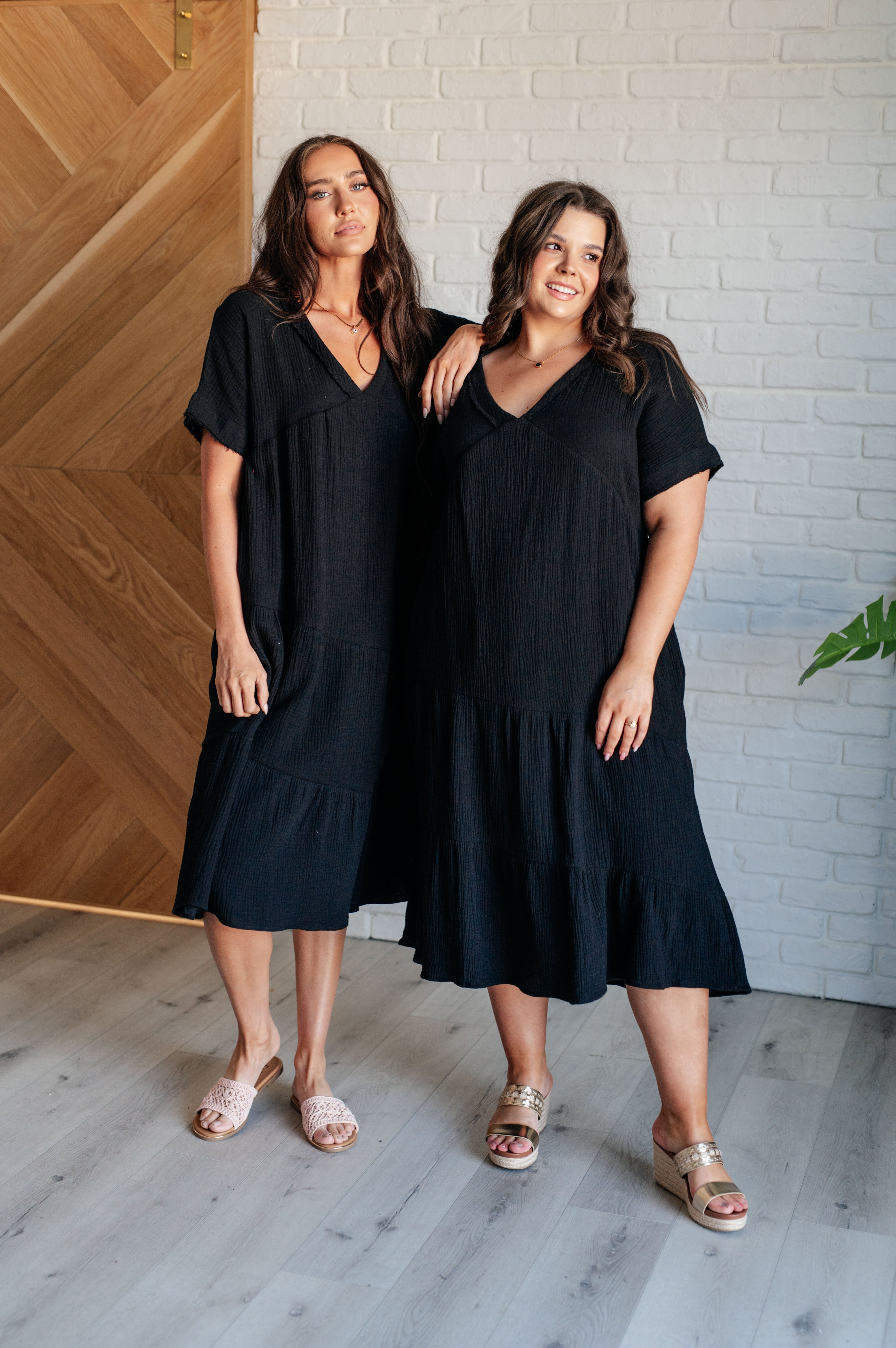 Two women standing in a room with wooden flooring and walls, wearing black dolman sleeve dresses with a V-neckline and tiered skirt.