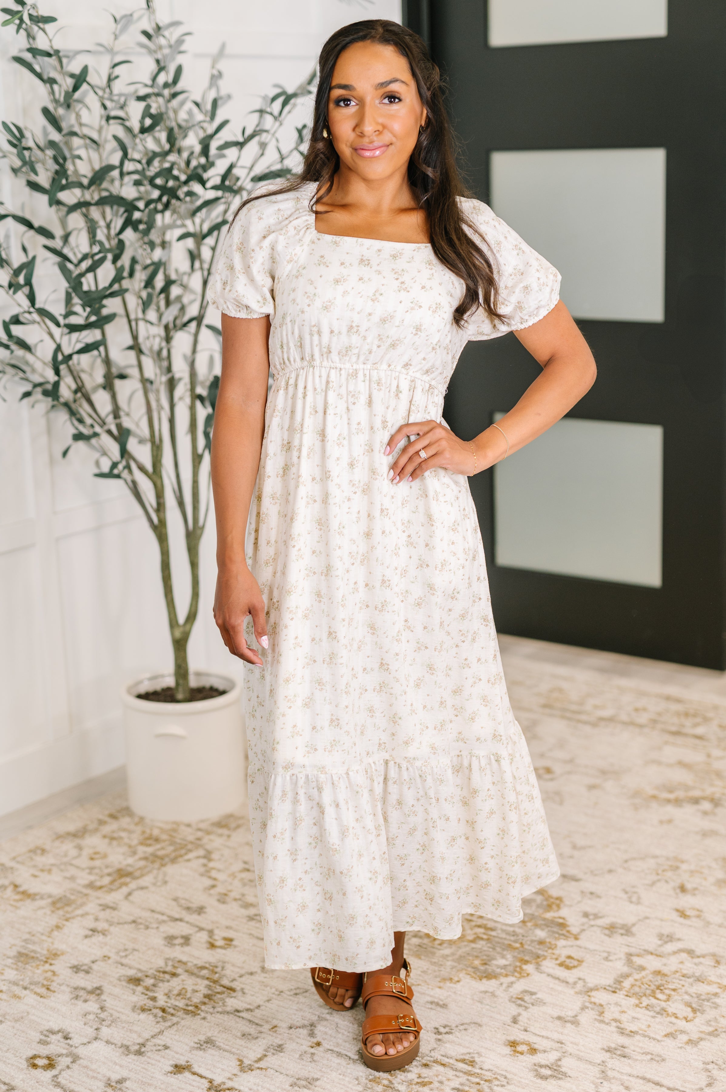 Woman wearing a white floral dress standing indoors with a plant and door in the background