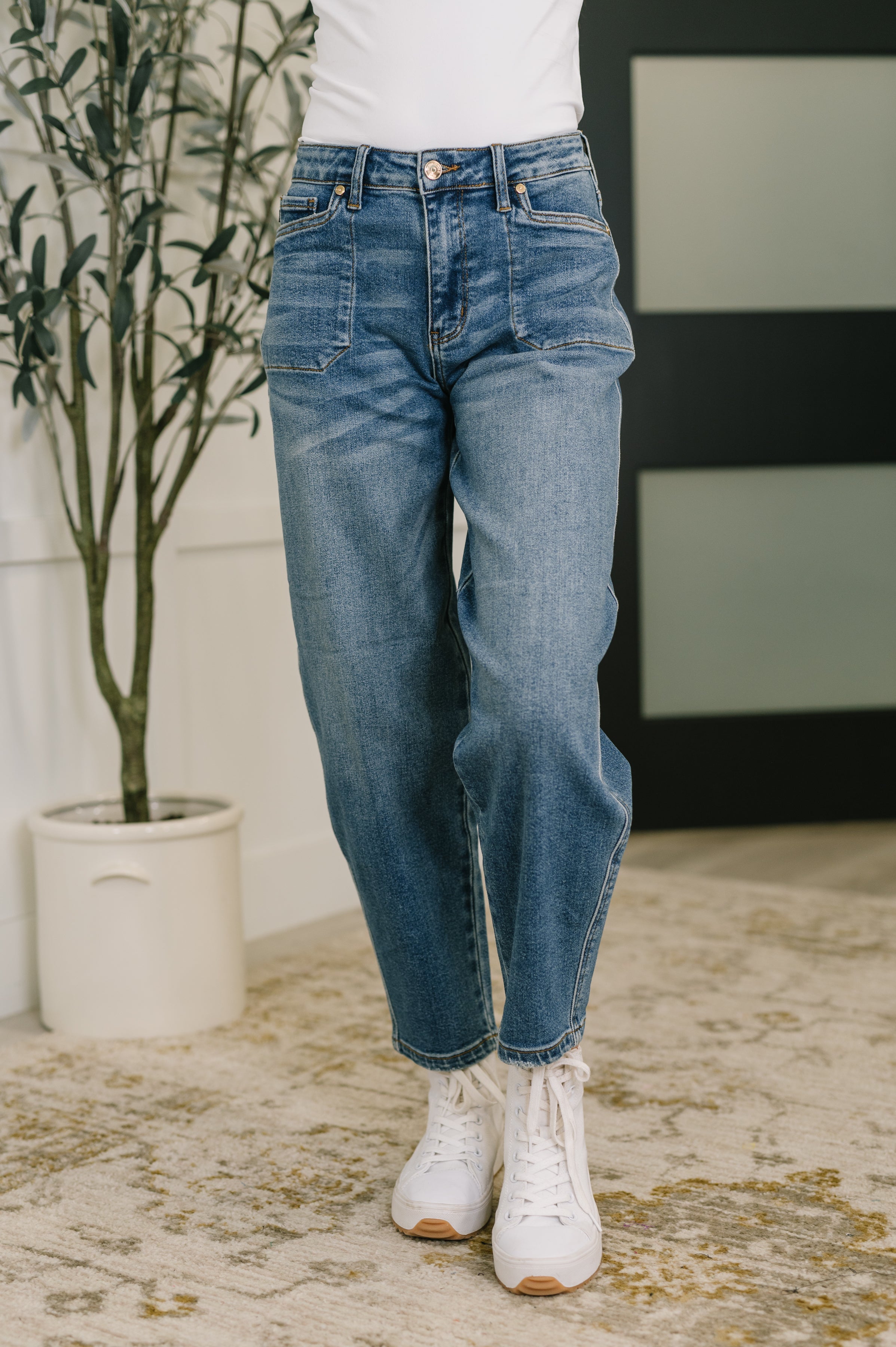 Woman wearing blue jeans and white sneakers standing in a room with a plant and striped wall.