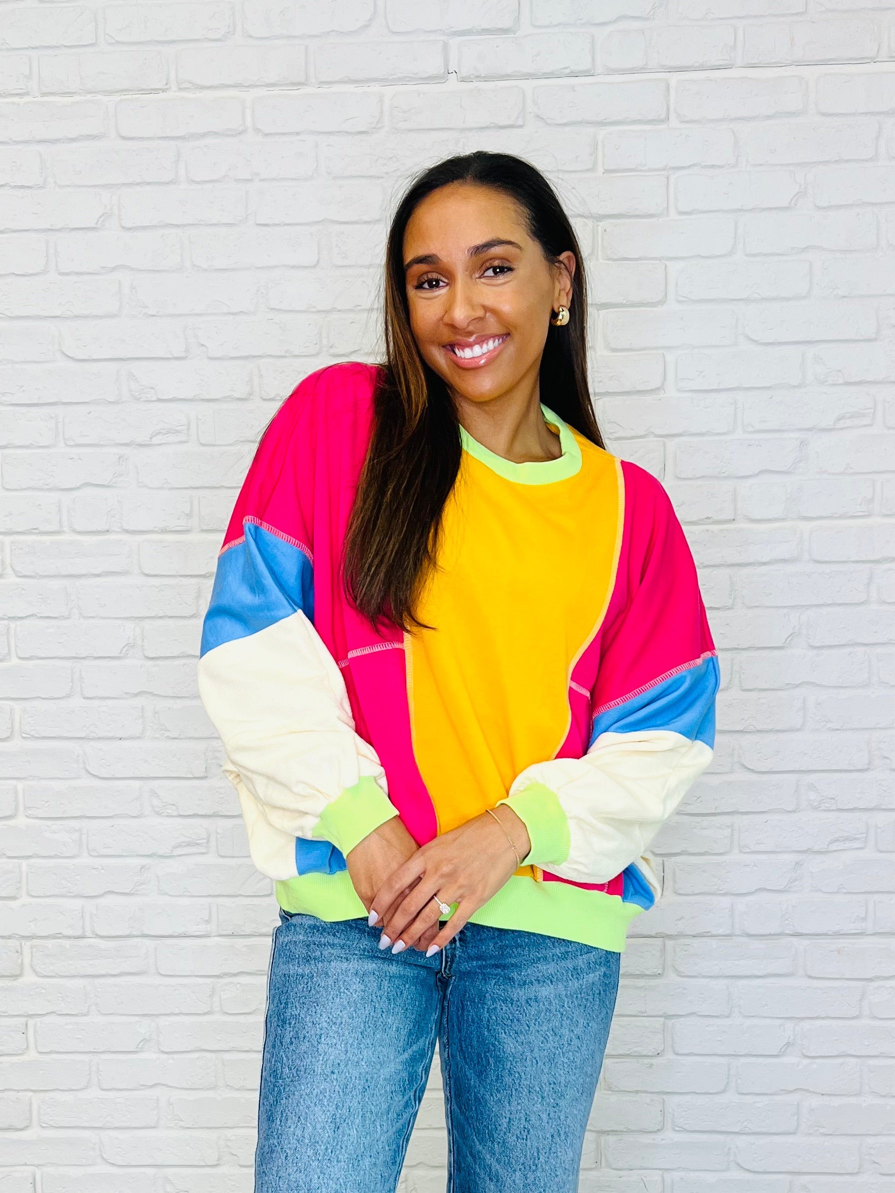 A woman wearing a multicolored block pullover with dolman sleeves and a ribbed neckline, paired with jeans, standing against a white brick background.