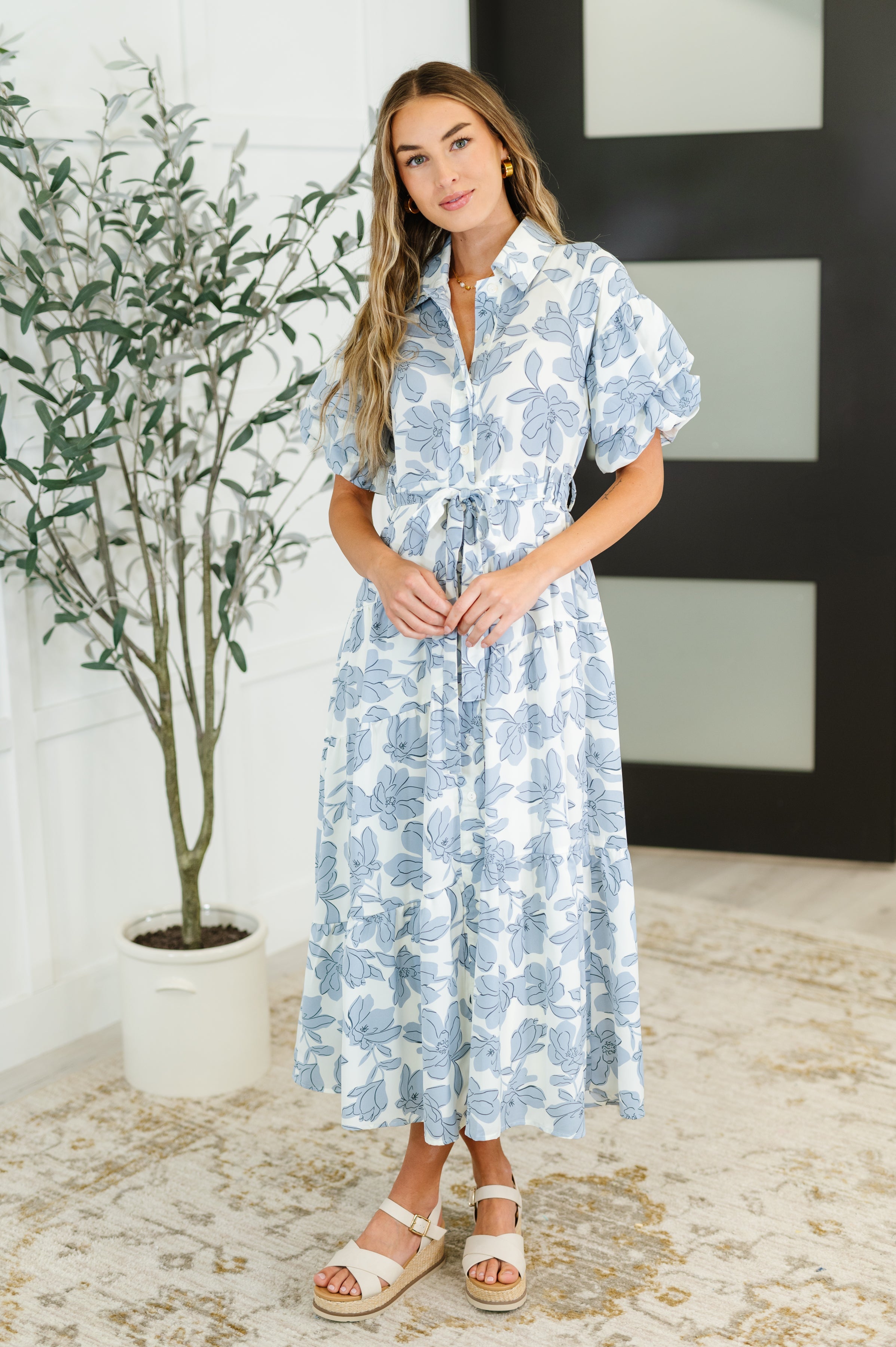 Woman wearing a blue floral dress standing indoors with a plant in the background