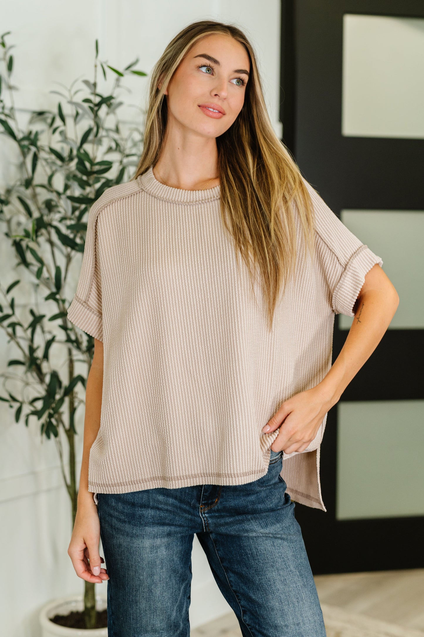 Woman wearing a beige short sleeve sweater and blue jeans standing indoors with a plant in the background