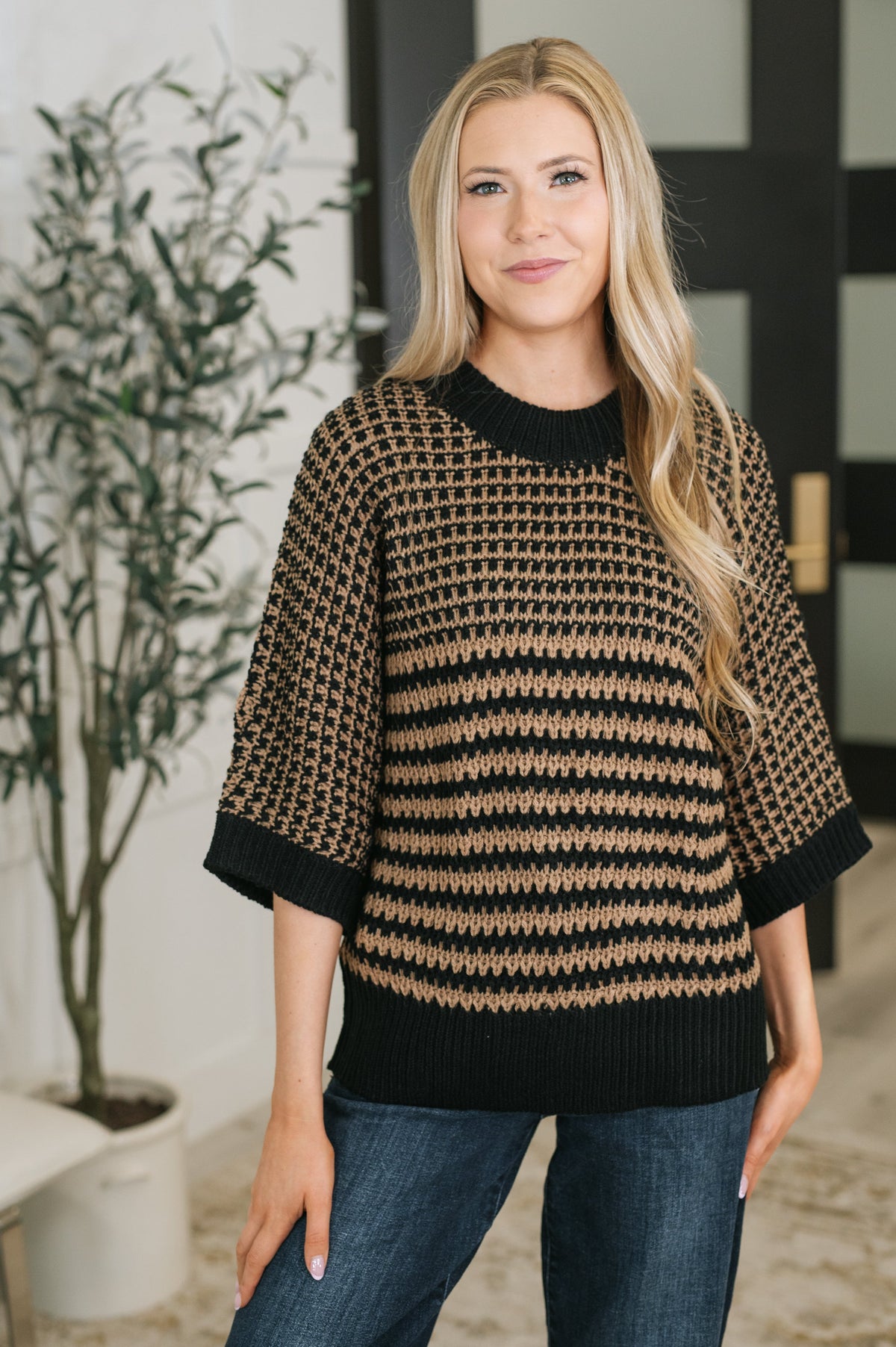 A woman wearing a black and tan knit sweater with a banded ribbed round neckline and dolman sleeves.