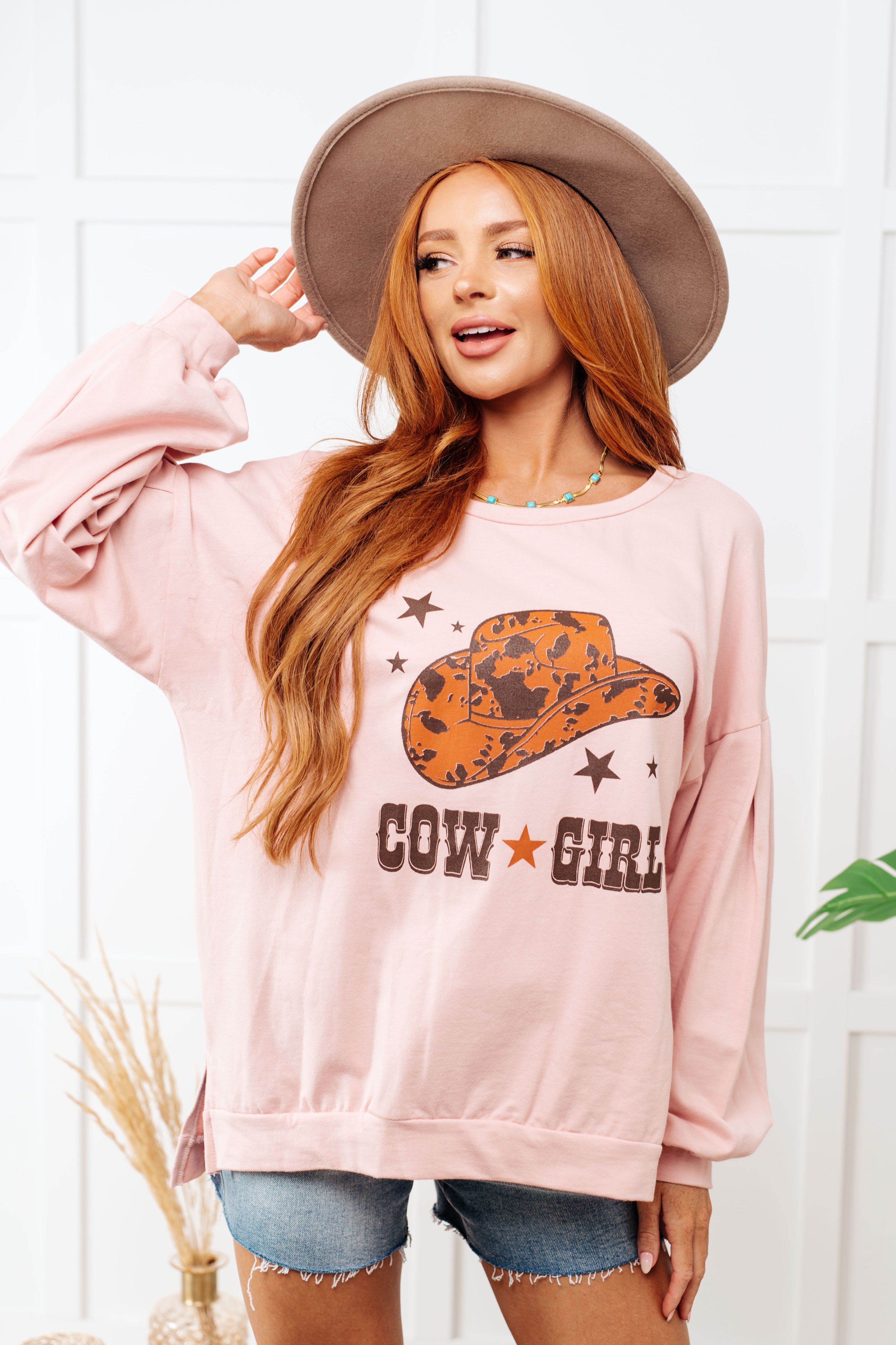 Woman wearing a pink sweatshirt with a cowgirl design and text, standing against a white wall.