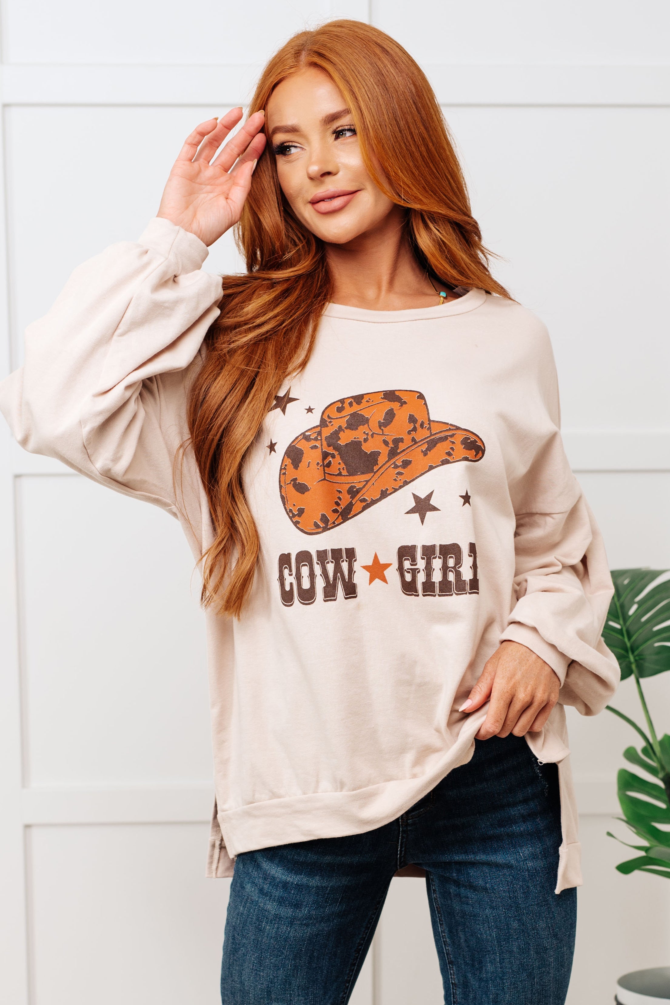 Woman wearing a beige sweatshirt with a cowgirl graphic and text, standing against a white wall.