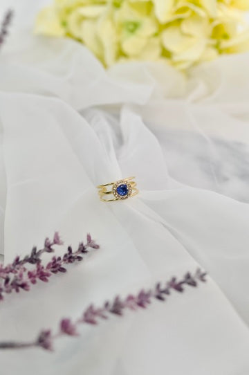 Gold tone open band ring with a blue cubic zircon stone on a soft white fabric surface.
