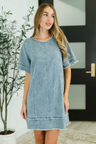 Woman wearing a light blue denim dress standing indoors with a plant and door in the background.