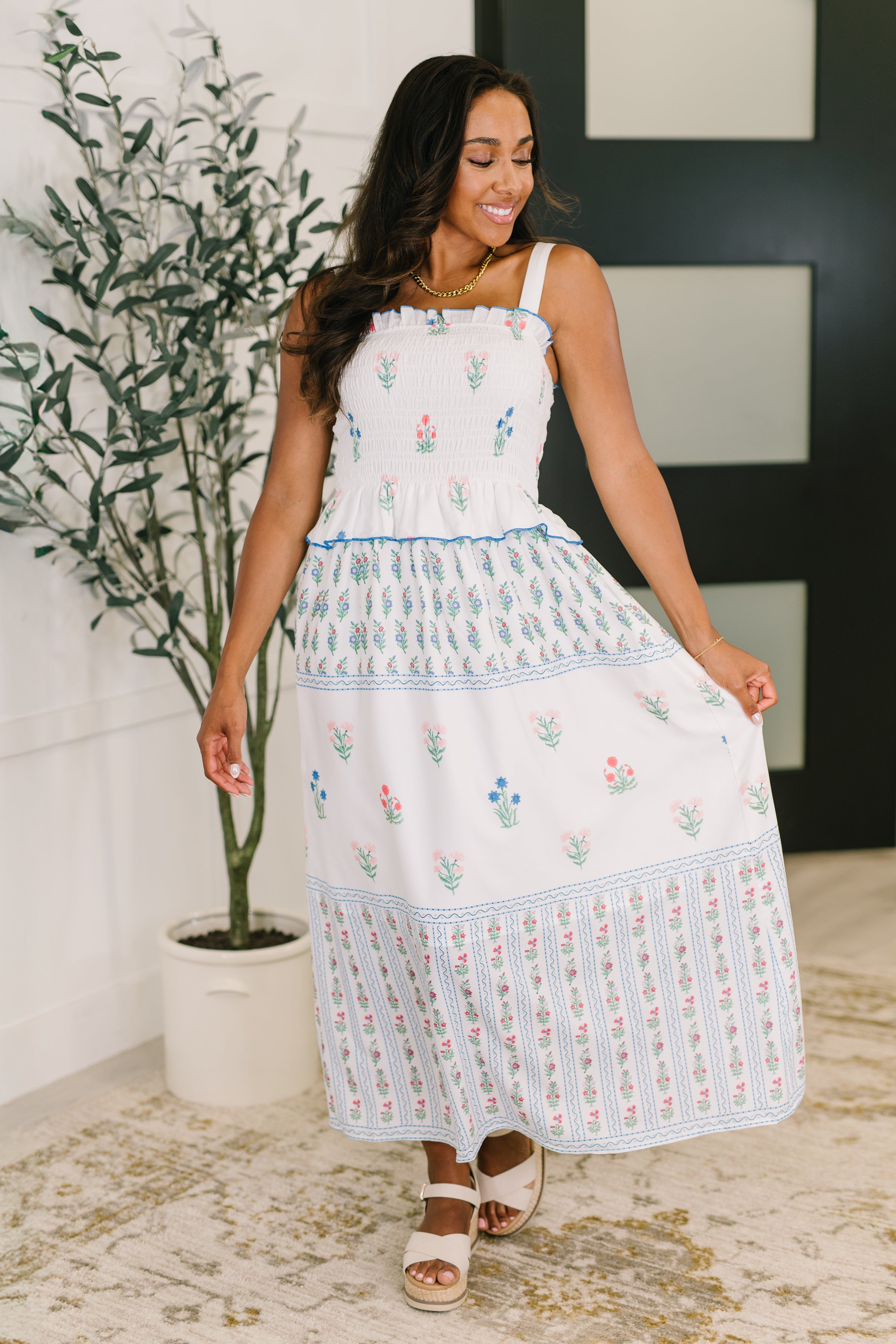 Woman wearing a white dress with floral patterns standing indoors next to a plant.