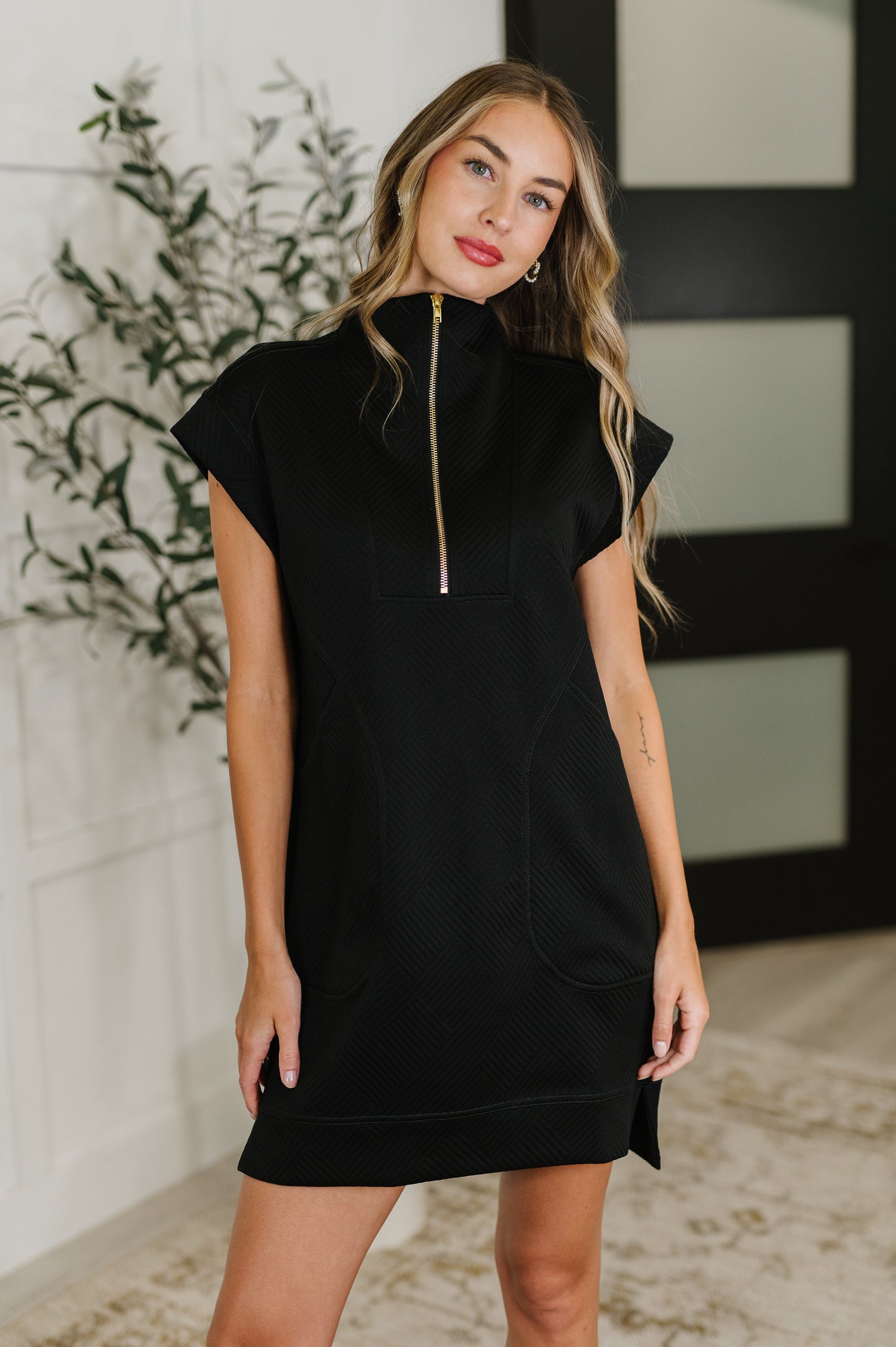 Woman wearing a black dress with a gold zipper in a room with decorative elements.