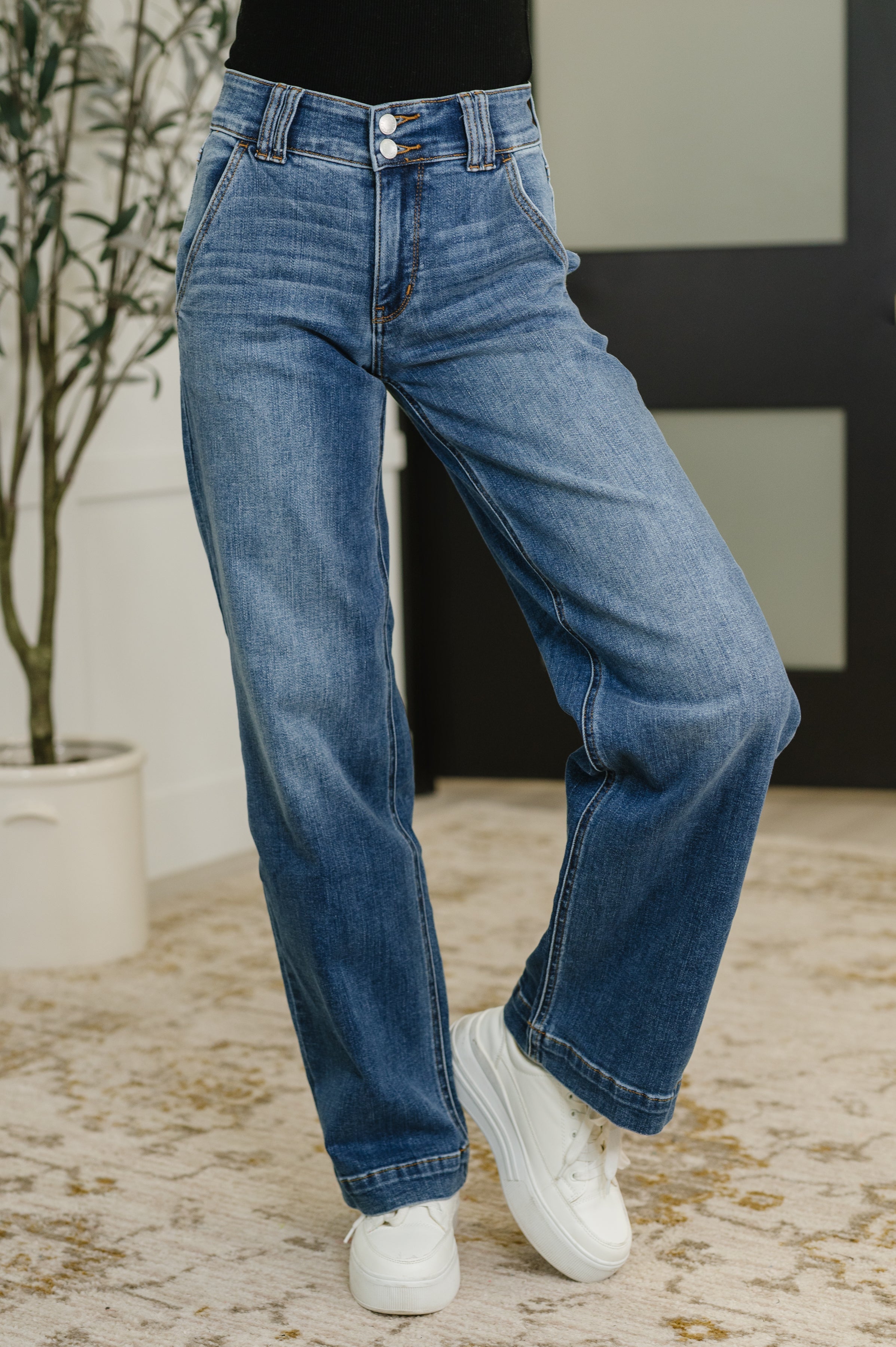Woman wearing blue jeans and white sneakers in a room with a plant and wall.