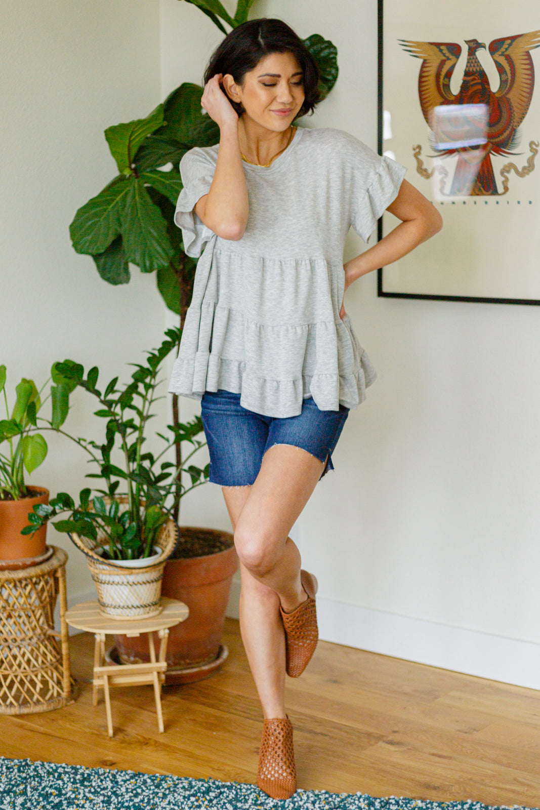 Woman in a gray ruffled top and blue shorts standing in a room with plants and a framed picture on the wall.