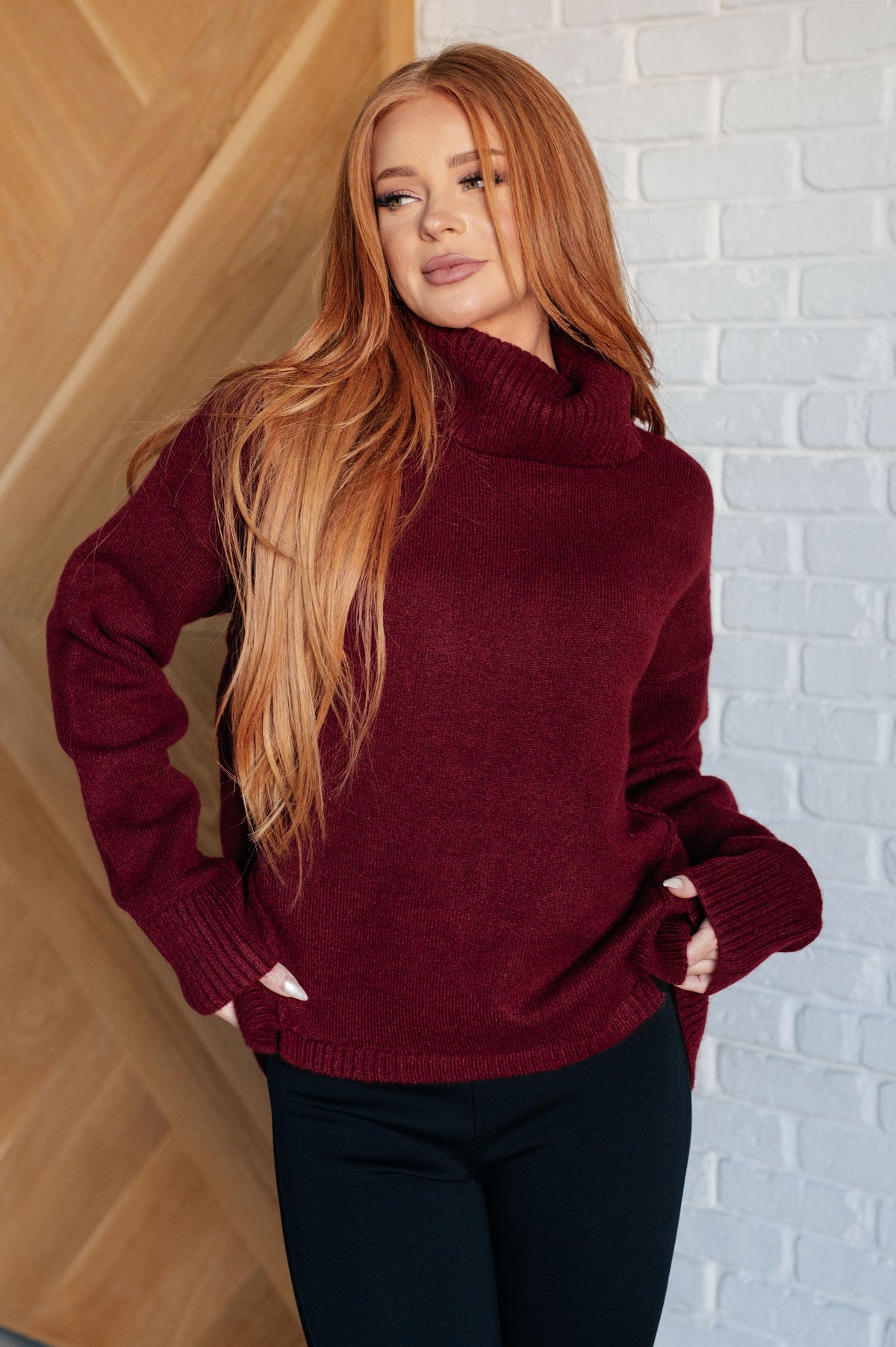 Woman wearing a burgundy turtleneck sweater with ribbed cuffs and a split stepped hem.