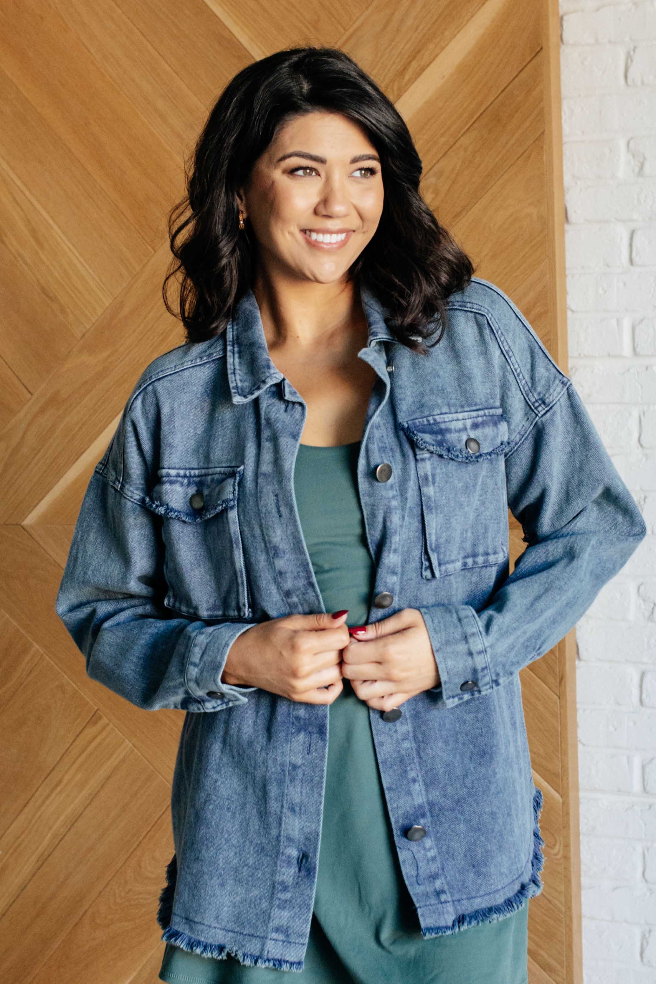 Woman wearing a denim shirt jacket standing against a wooden paneled wall.