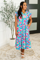 Woman wearing a blue floral dress standing indoors with a plant in the background