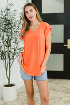 Woman wearing an orange top and denim shorts standing indoors next to a plant.