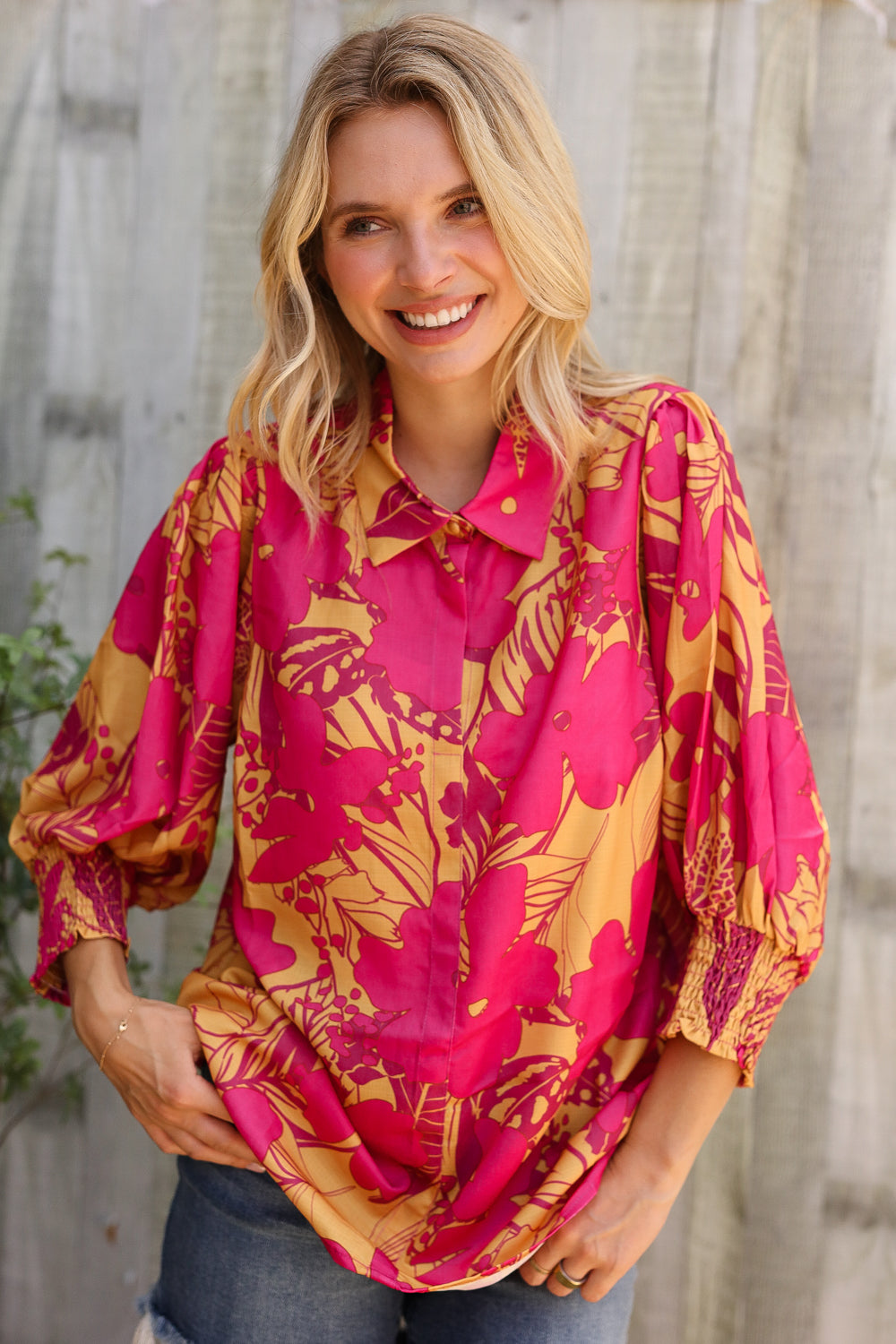 Woman wearing a pink and yellow floral blouse against a wooden fence background