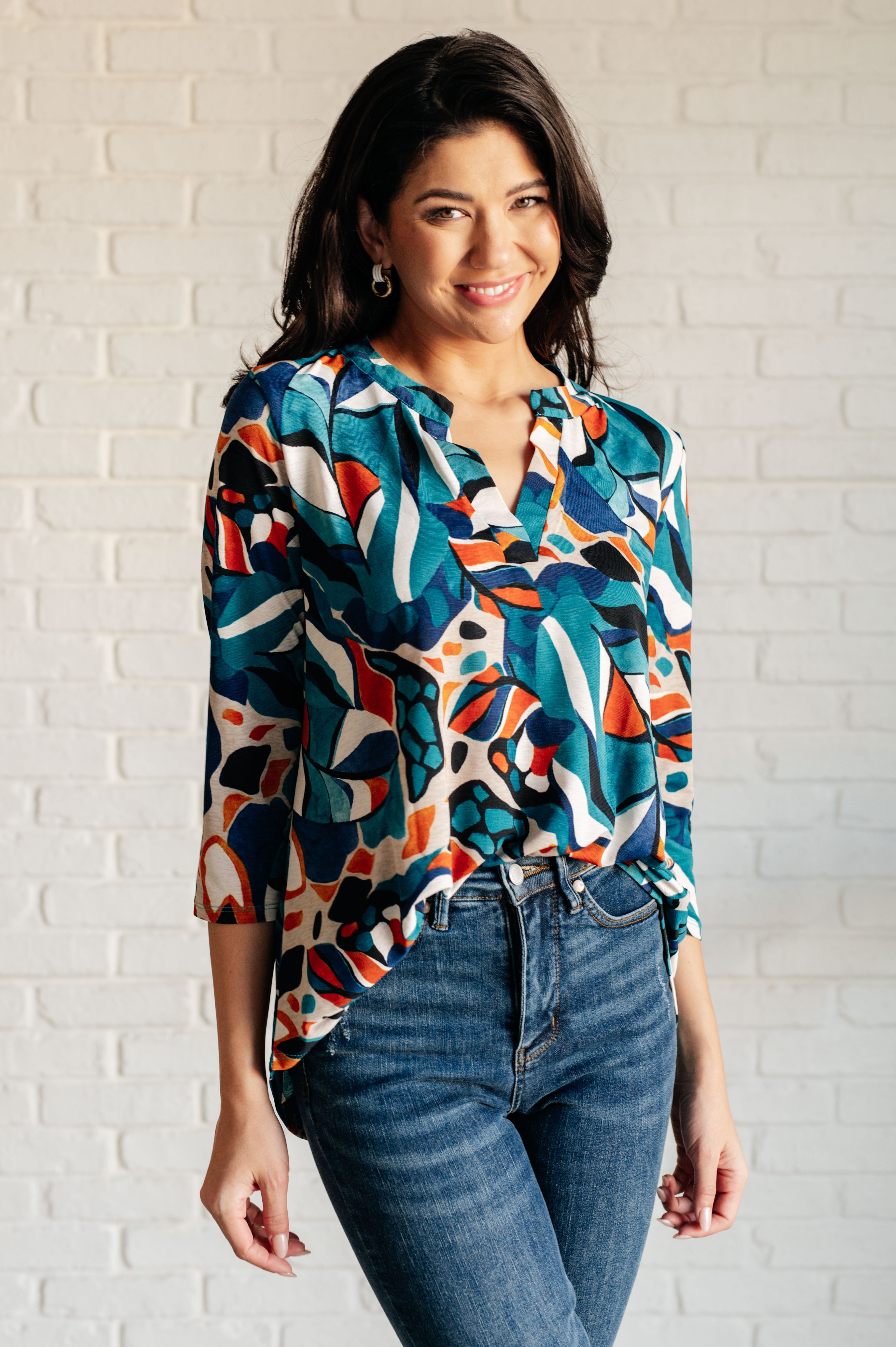 Woman wearing a colorful patterned blouse and jeans against a white brick wall.