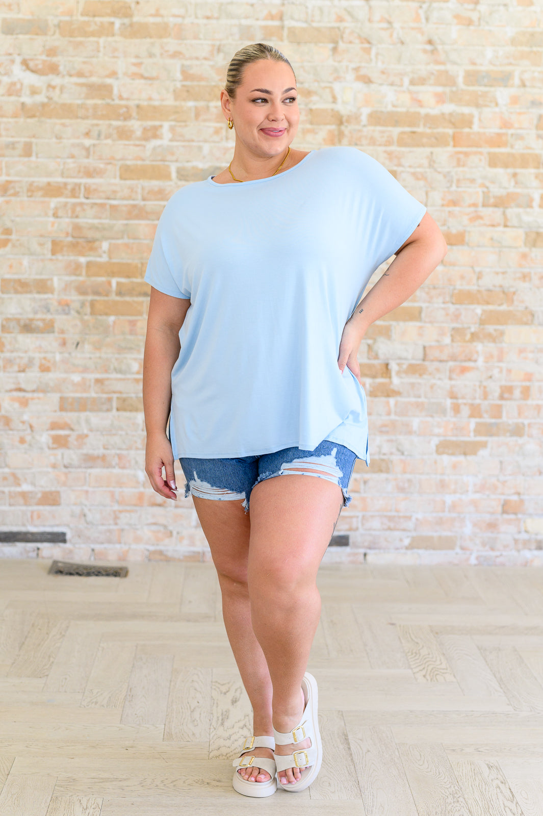 A woman wearing a light blue, round neck, dolman sleeve top with side slits paired with denim shorts and white shoes.