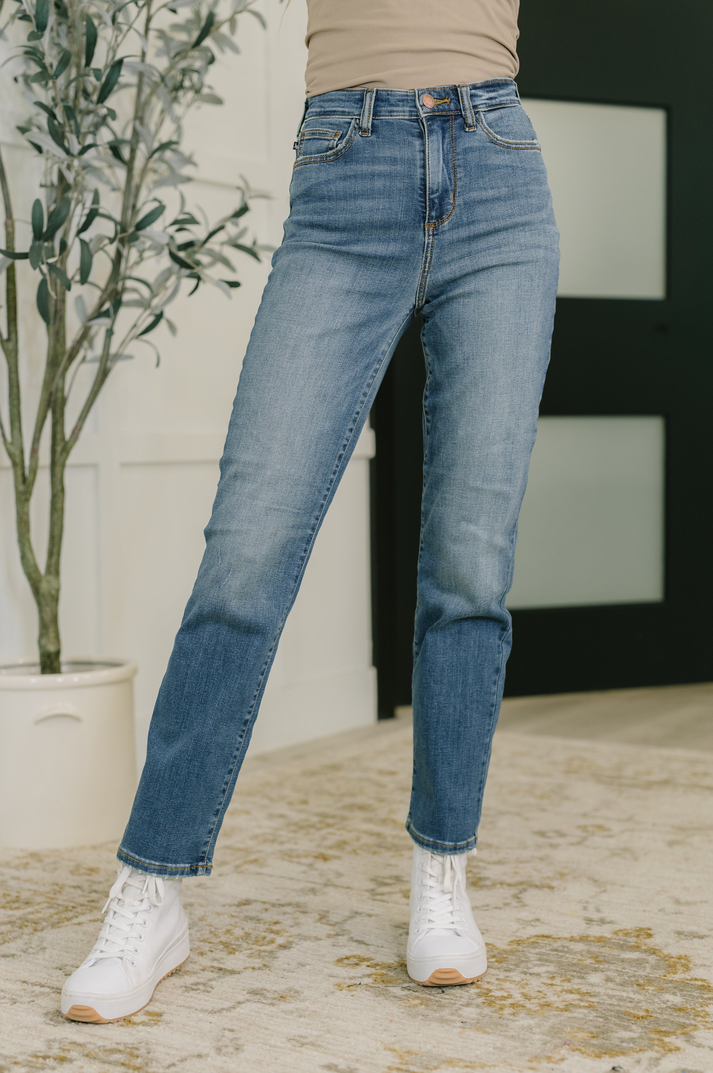 Woman wearing blue jeans and white sneakers standing indoors with a plant in the background