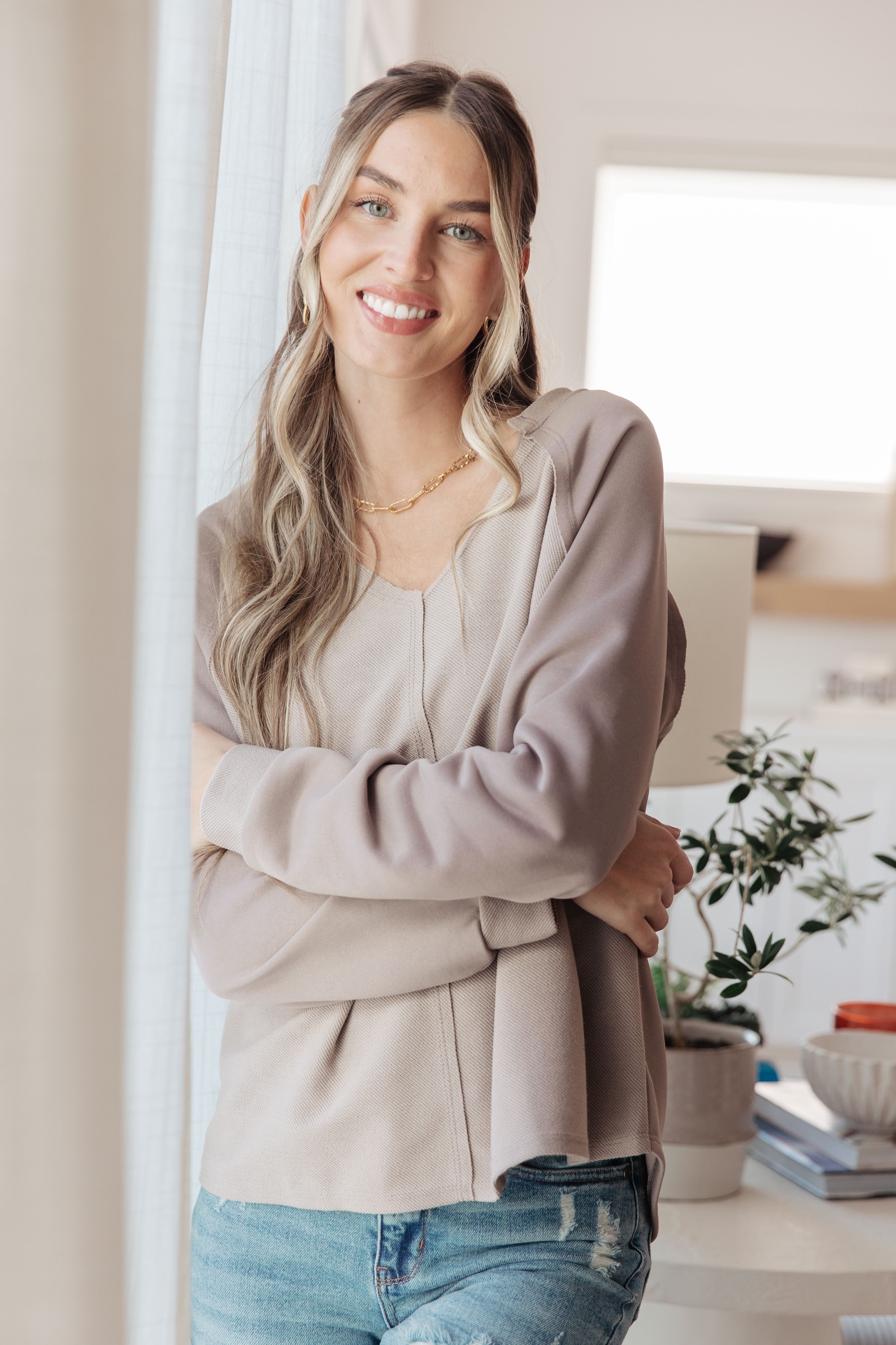 Woman standing indoors wearing a light gray shirt top and blue jeans, smiling.