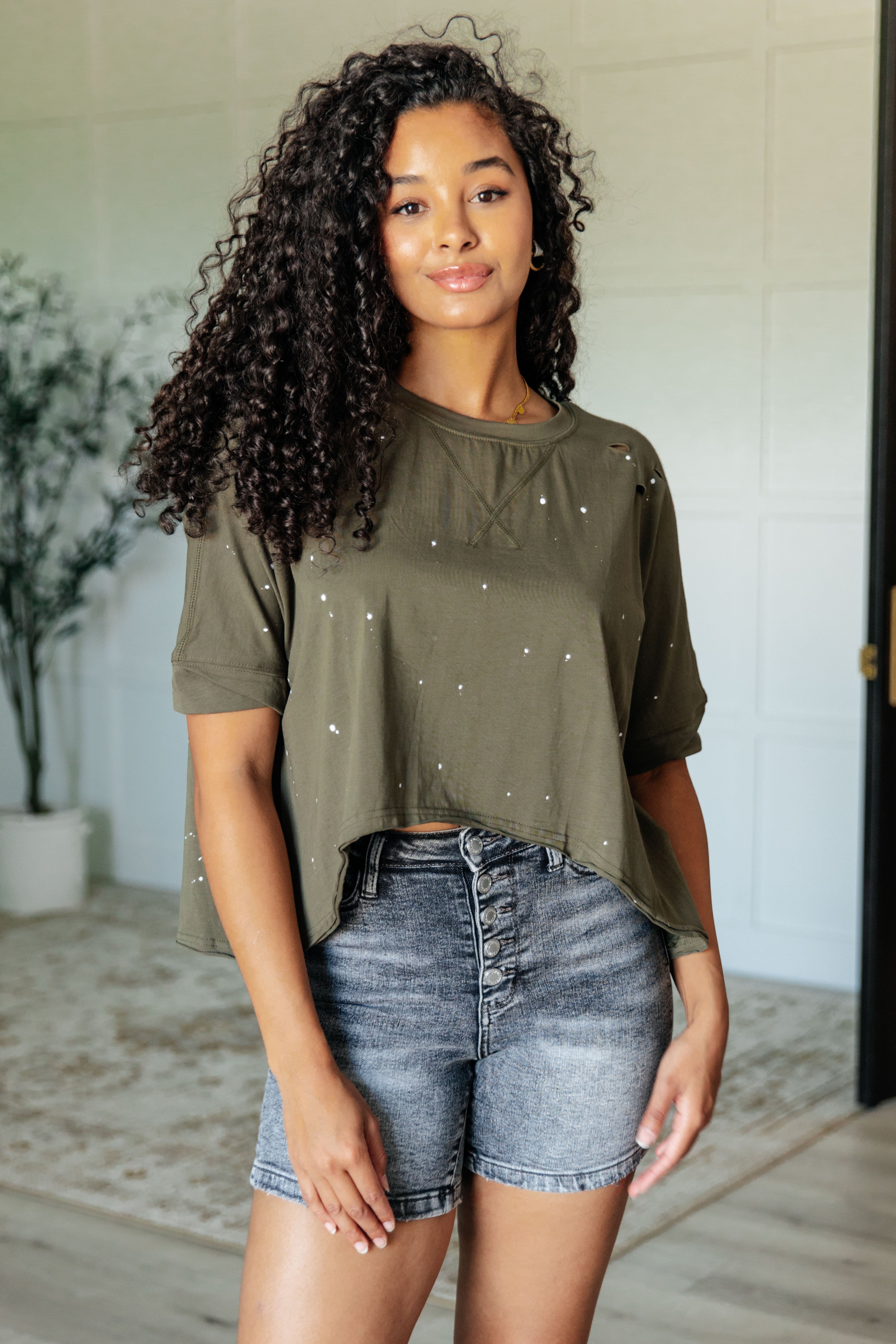 A woman wearing an olive green asymmetrical distressed top with a round neckline and paint splatter details, paired with denim shorts.