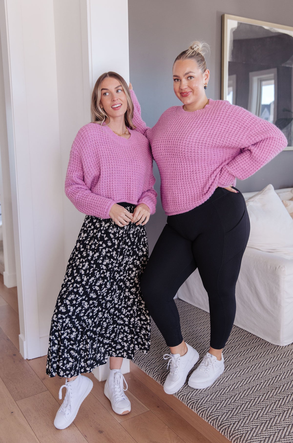 Two models wearing pink waffle knit sweaters, one with a floral skirt and the other with black leggings.