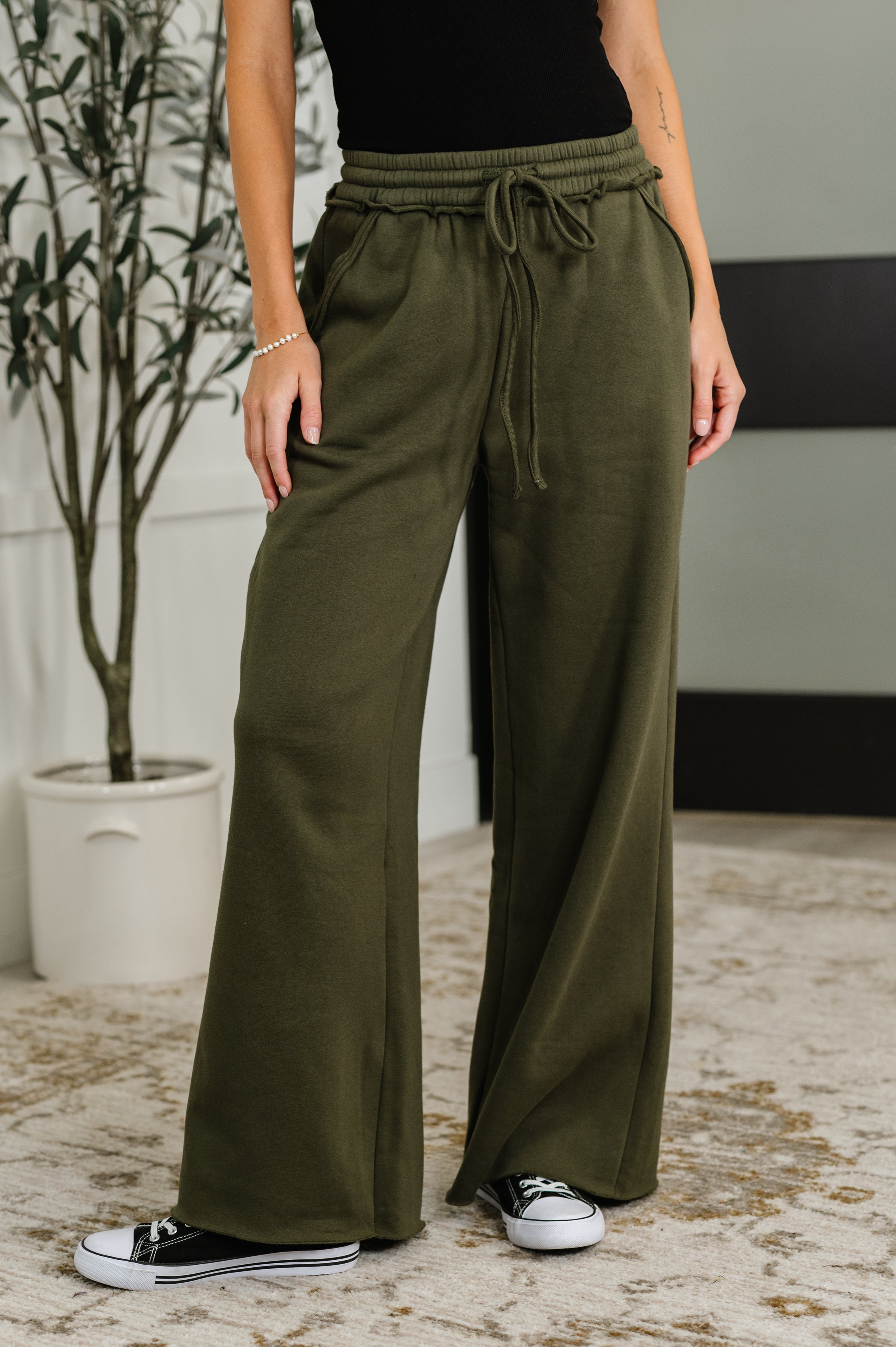 Woman wearing olive green wide-leg pants with a plant and wall in the background