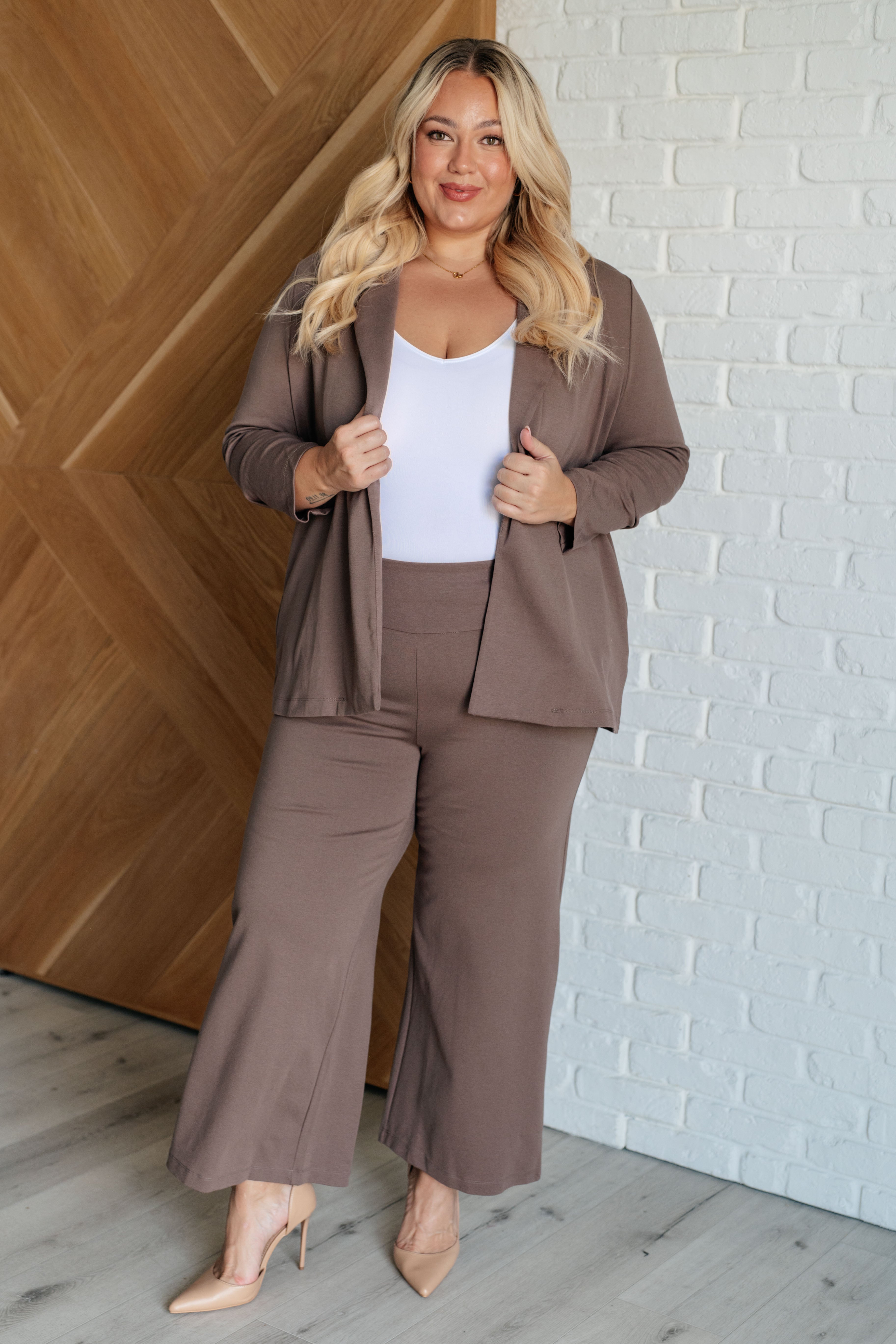 A woman standing in a doorway wearing a dark mocha colored blazer with ruched sleeves and a notched lapel, paired with a white top and beige trousers.