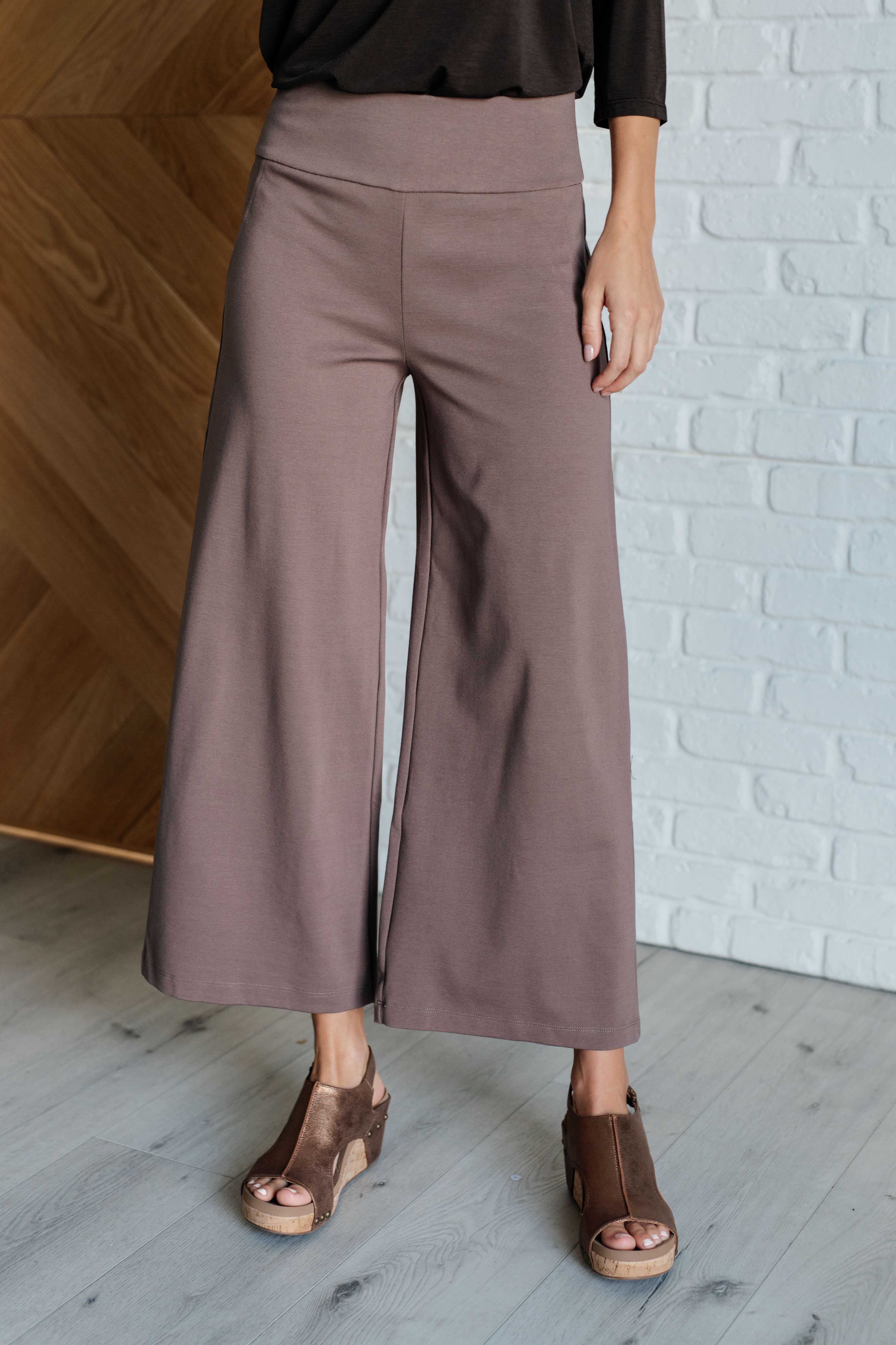 A woman wearing dark mocha wide leg crop pants with a faux welt pocket, standing in a room with wooden flooring and a white brick wall.