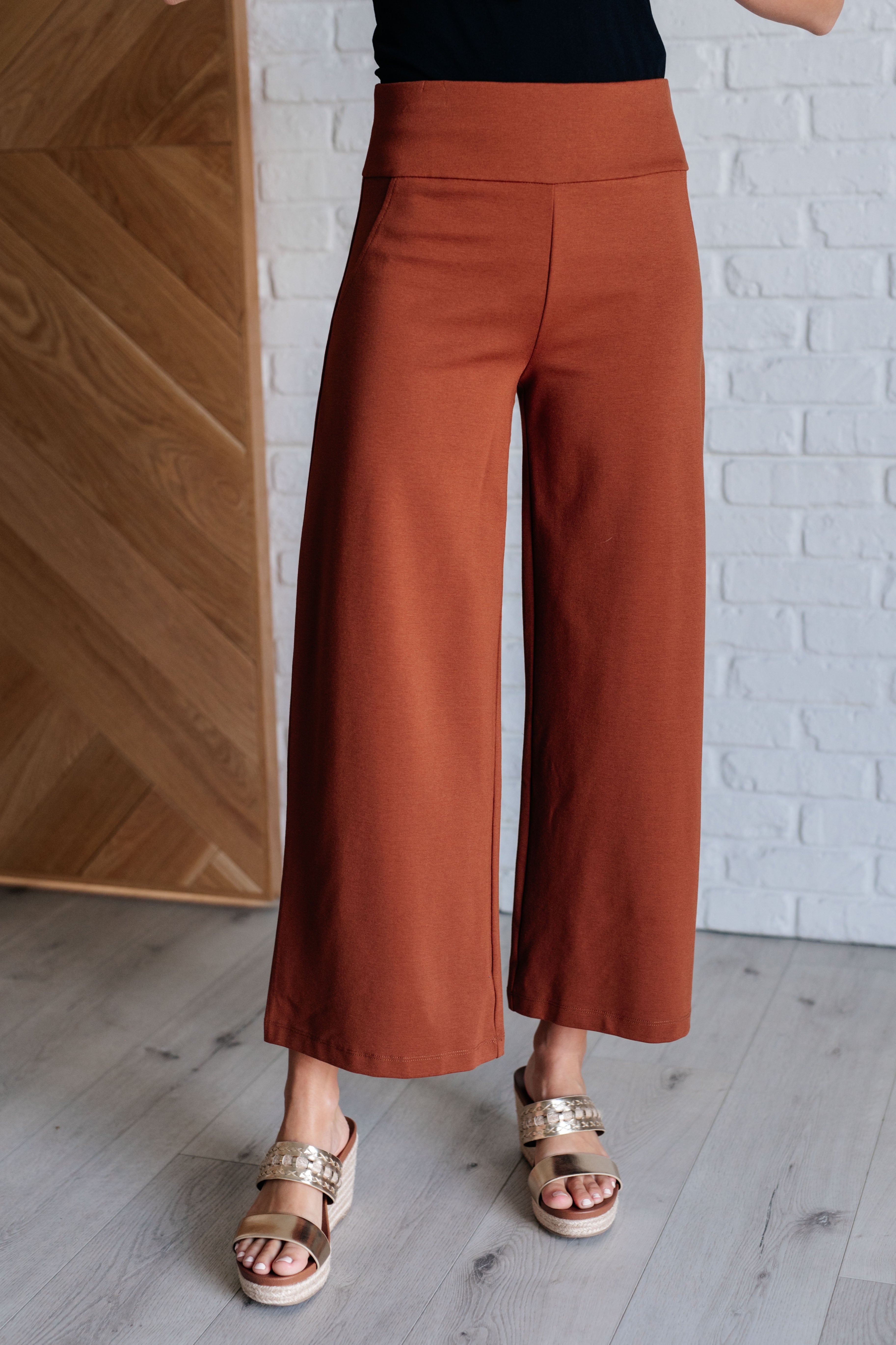 A pair of rust-colored wide leg crop pants with a faux welt pocket, displayed on a model wearing open-toe sandals.