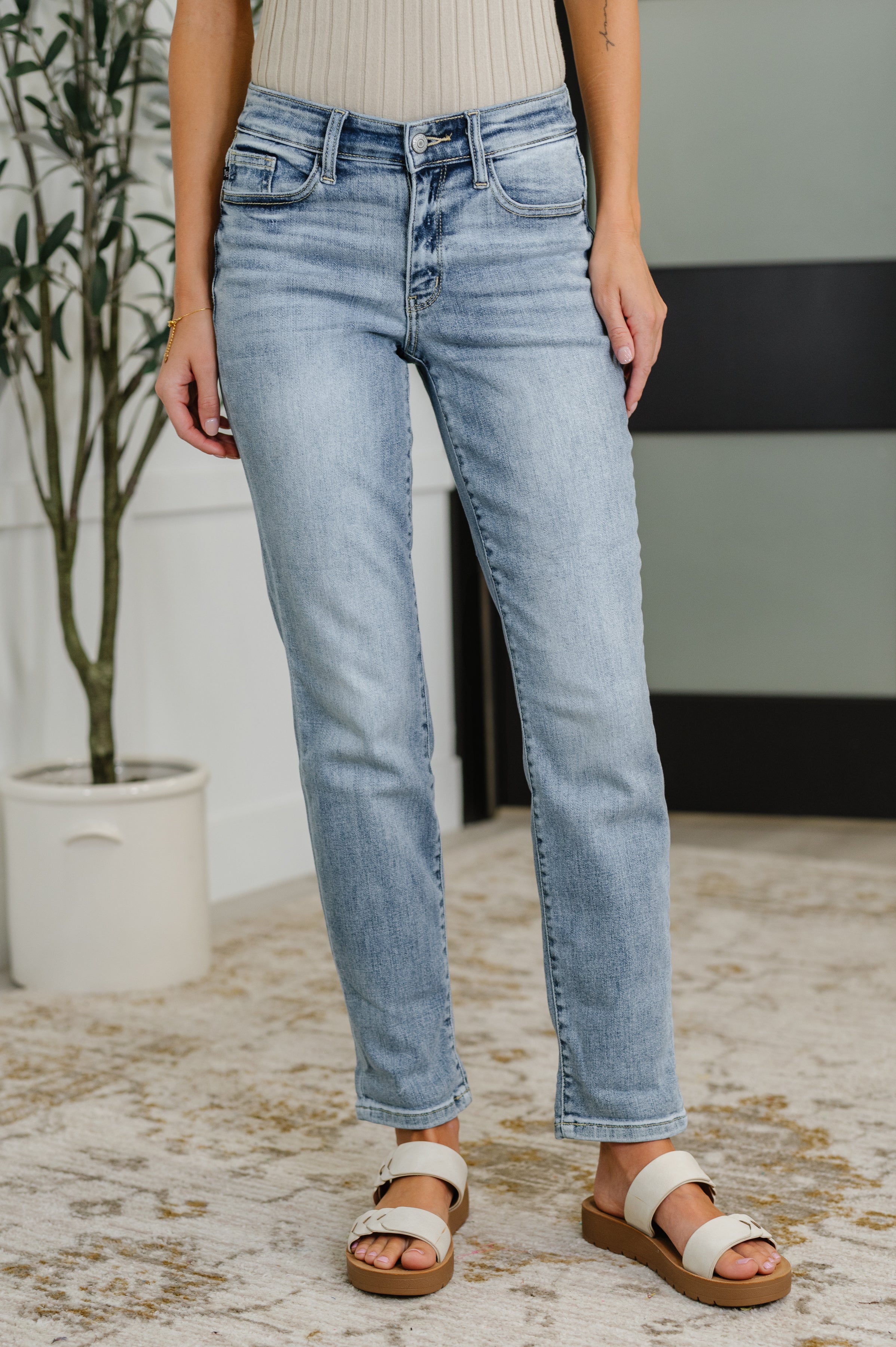 Woman wearing light blue jeans standing indoors with a plant and wall in the background