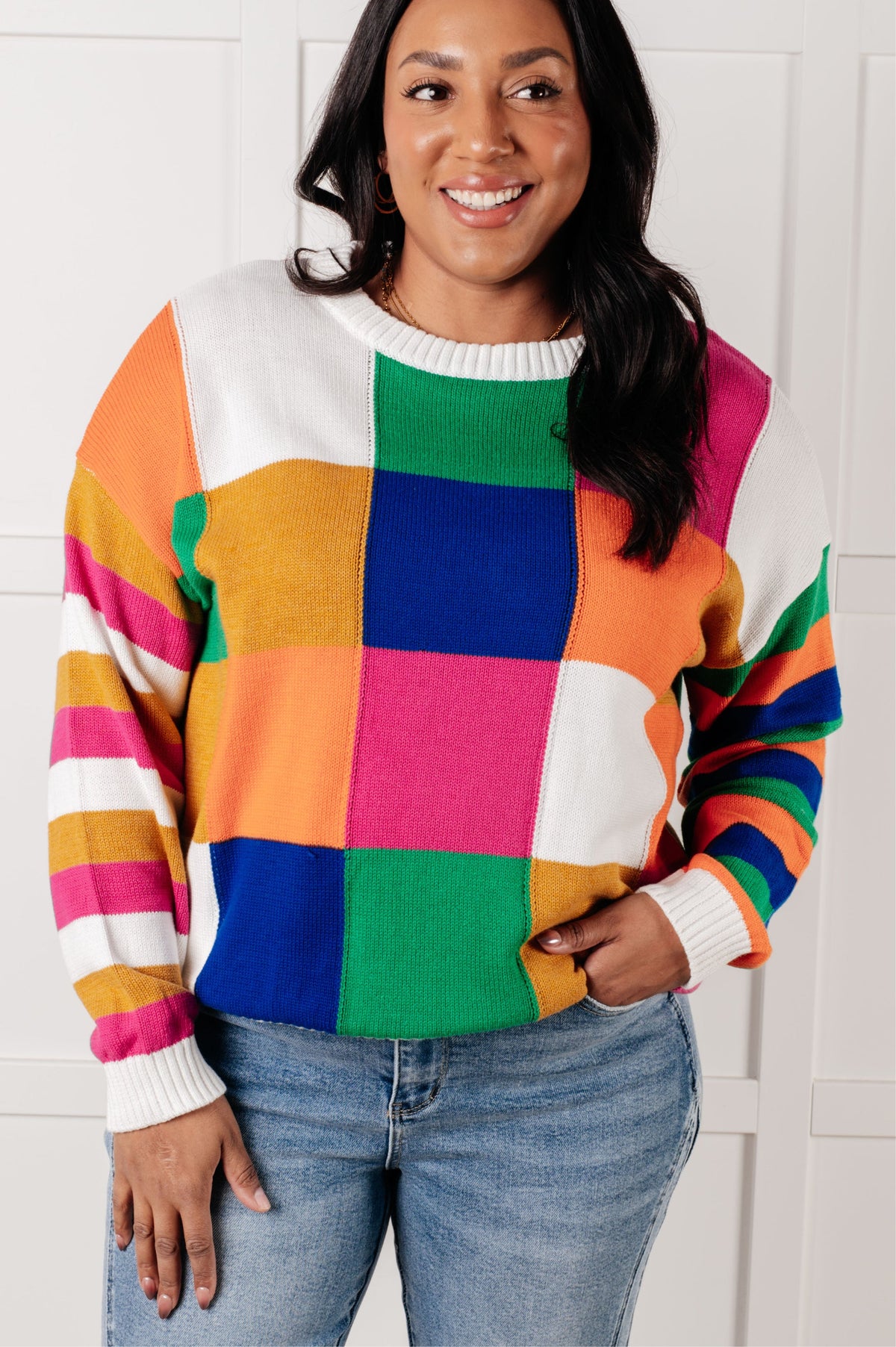 A colorful checkered color block sweater with a ribbed round neckline and drop shoulders, modeled with jeans.