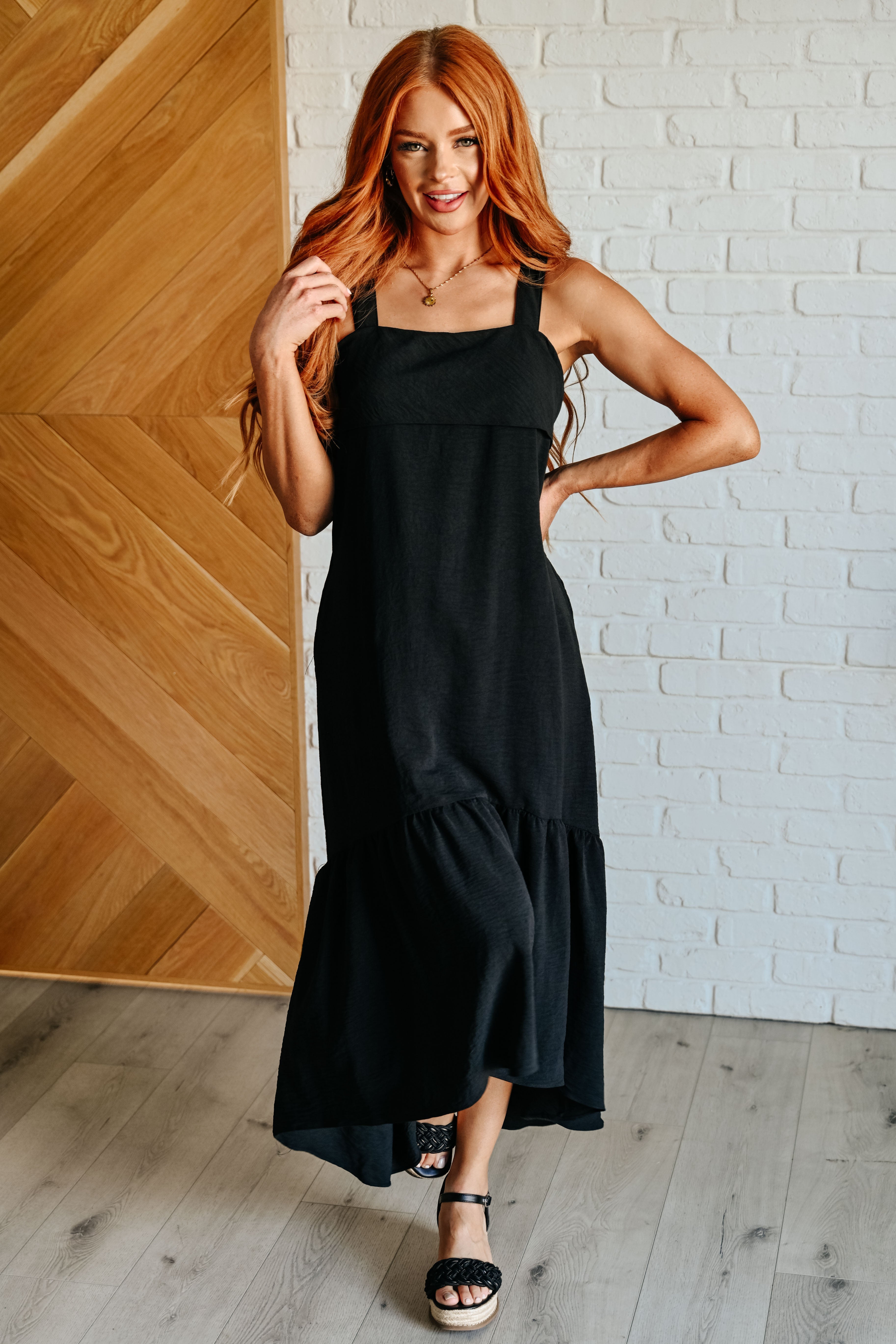 A woman wearing a black tie back maxi dress with a high low hem and side pockets, paired with heels.