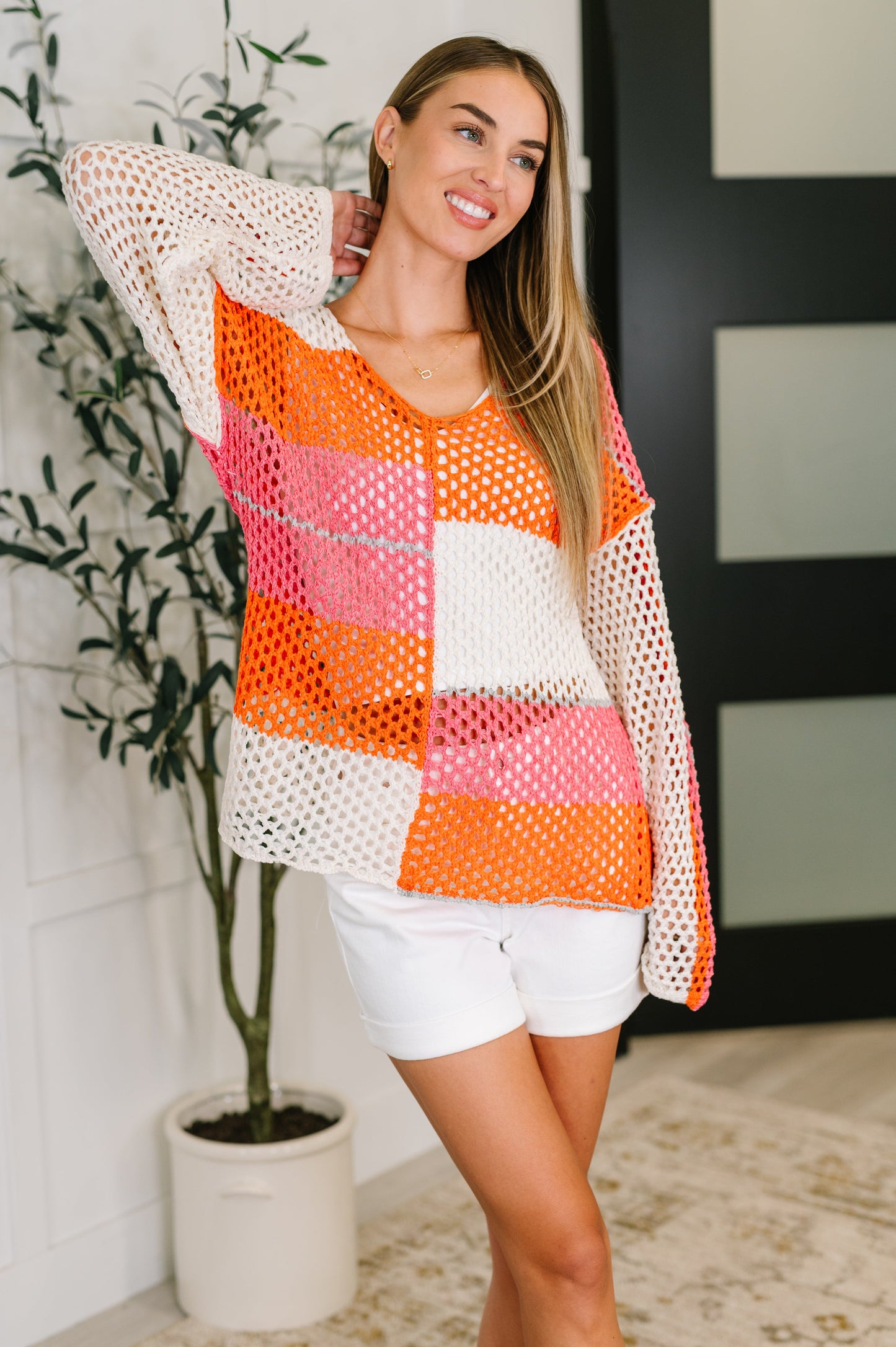 Woman wearing a colorful crochet top and white shorts standing indoors with a plant in the background.