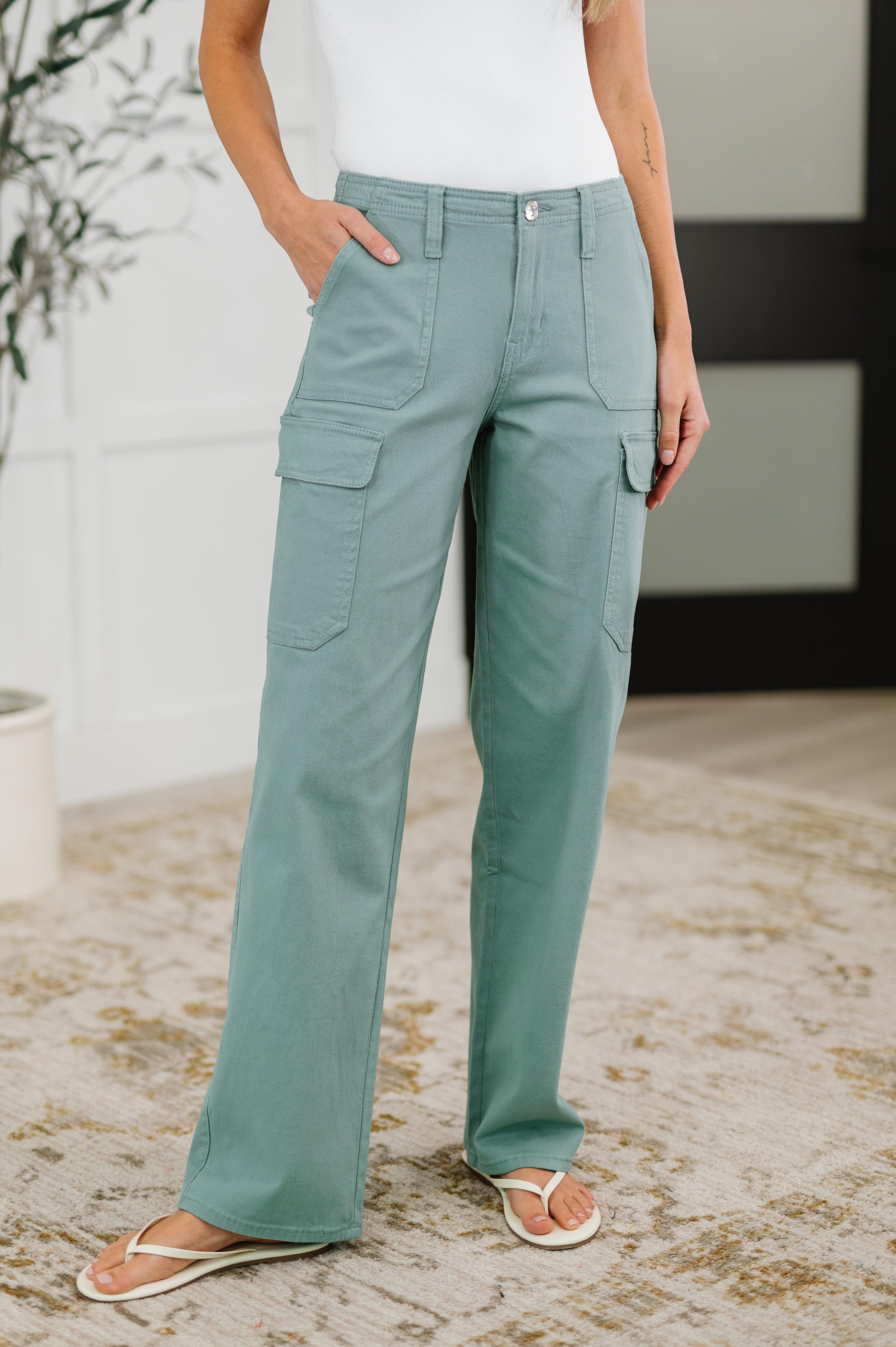 Woman wearing mid-rise, straight-leg cargo bottoms in a solid green color with functional patch pockets, belt loops, and a zipper and button closure.
